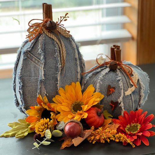 Two Eclectic Spruce decorative pumpkins made from faded blue denim fabric with rustic fringe, faux cinnamon sticks, ribbon, and twine. They are styled with autumn flowers and leaves on a dark tabletop in front of a window.