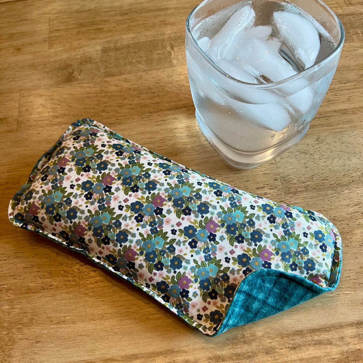 An Eclectic Spruce Wildflower Glen Weighted Eye Pillow on a wood surface next to a glass of ice water.