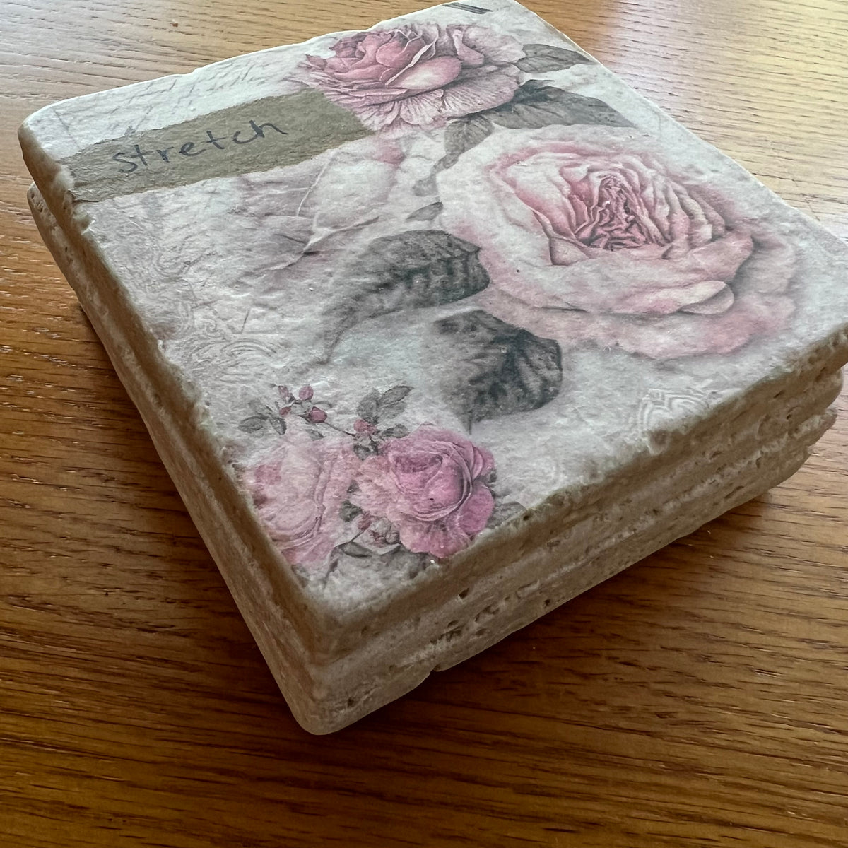 A set of four travertine coasters by Eclectic Spruce stacked on top of one another on a wooden surface. The coasters feature a vintage floral design with pink roses and dark leaves along with a small brown paper label that reads 'stretch'.