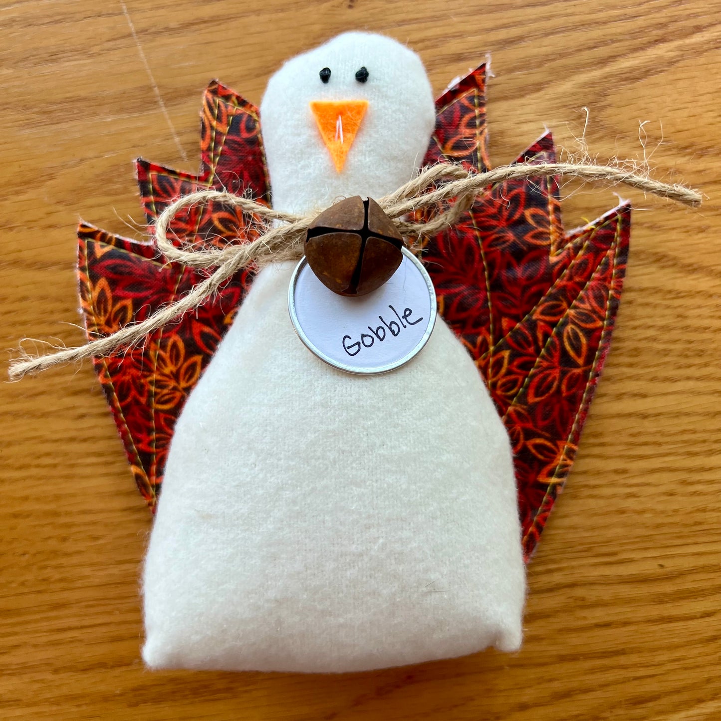An Eclectic Spruce handmade fabric Thanksgiving turkey sitting on a wooden table. The turkey features a white flannel body, textile tail feathers, an orange felt beak, black stitched eyes, a brown rusty bell on a twine bow, and a round white tag that reads "Gobble."