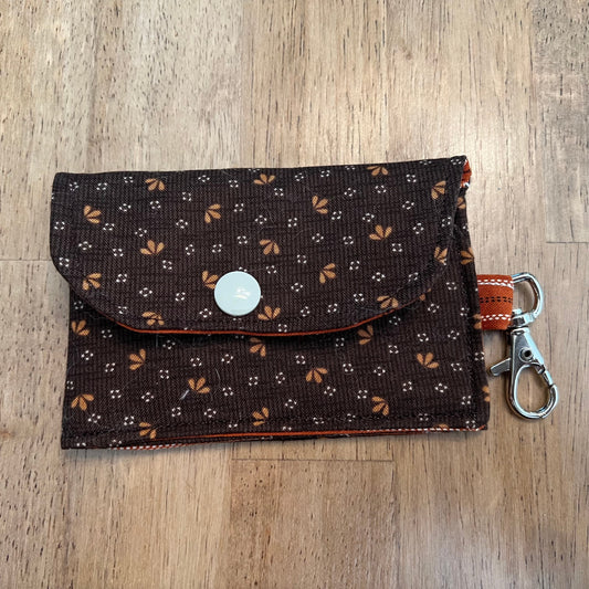 Eclectic Spruce handcrafted mini card holder wallet keychain with a snap button closure. The wallet has a dark brown exterior fabric with a small-scale floral print in white and orange-brown and contains a matching orange-brown fabric loop and silver metal swivel clasp.