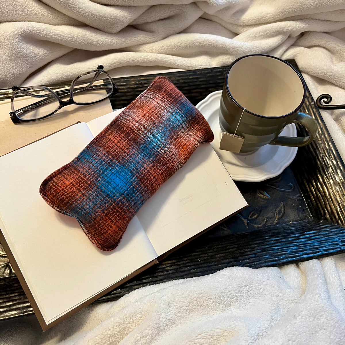 A Copper Ridge Eclectic Spruce Weighted Eye Pillow sitting on a tray with a book, mug, pair of glasses, and a cozy white blanket.