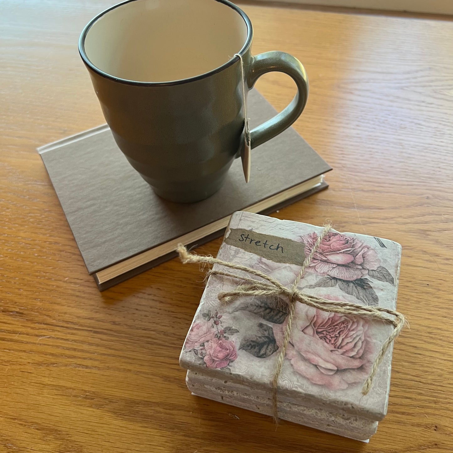 A styled tabletop scene with a gray ceramic mug resting on a brown hardcover book, alongside a set of four Eclectic Spruce travertine coasters stacked together and tied with rustic jute twine. The coasters feature a floral pink rose design and have a small label reading 'Stretch'.