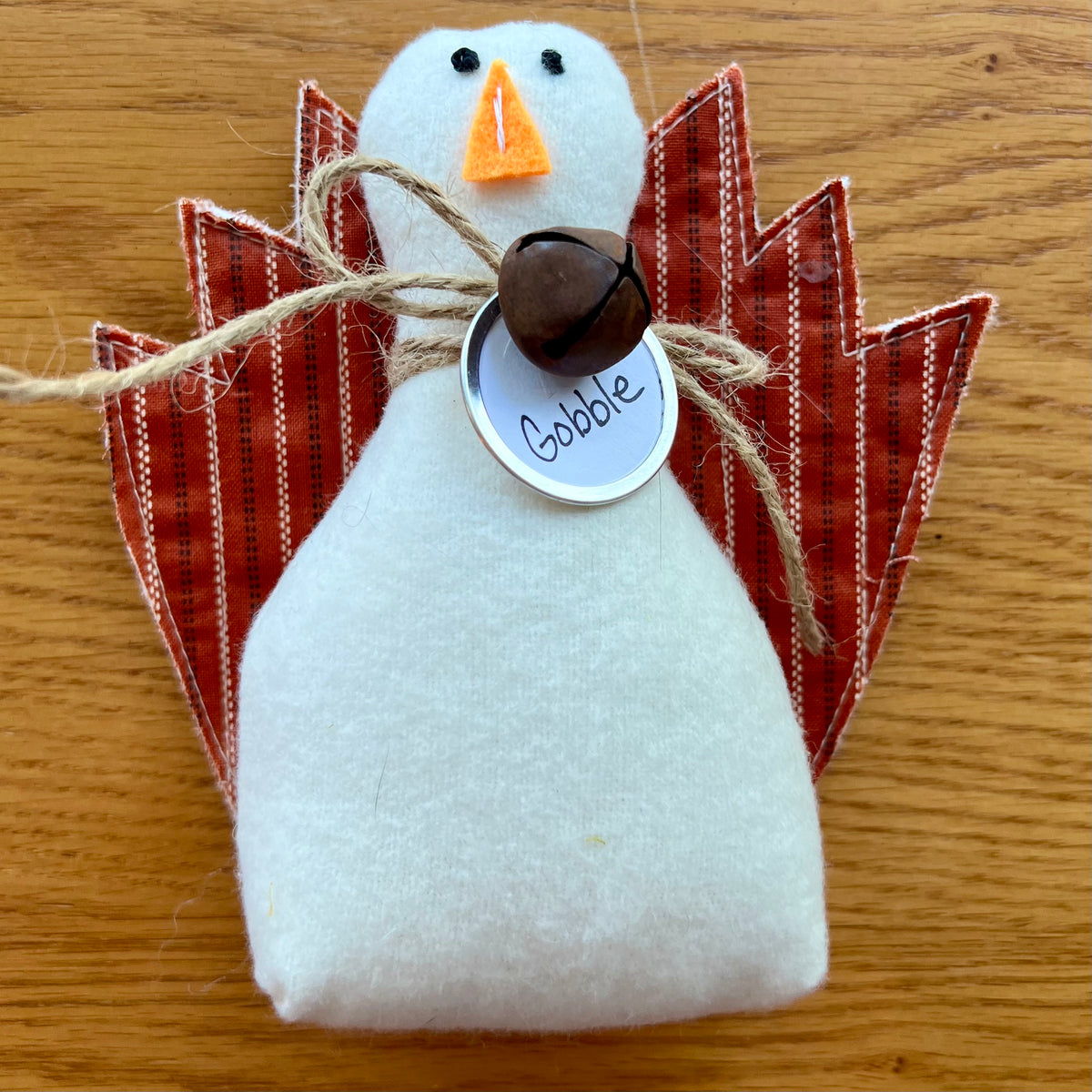 An Eclectic Spruce handmade fabric Thanksgiving turkey sitting on a wooden table. The turkey features a white flannel body, textile tail feathers, an orange felt beak, black stitched eyes, a brown rusty bell on a twine bow, and a round white tag that reads "Gobble."