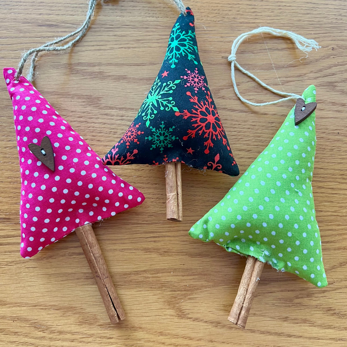 Eclectic Spruce's handmade cinnamon stick trees are made with cotton fabric, stuffed with fiberfill, and embellished with real cinnamon sticks along with lace, buttons, bells, and other trim.  Sold mystery style, you will receive a curated collection of 3 unique trees picked out just for  you. This photo shows 3 trees arranged on a wooden surface.