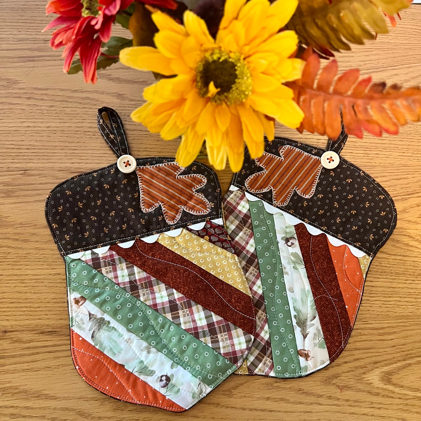 Two Eclectic Spruce acorn-shaped quilted potholders with autumn-themed patchwork fabrics, applique leaf design, wood buttons, rick rack detailing, and hanging loops. They are displayed on a wooden surface with decorative autumn colored flowers in the background.