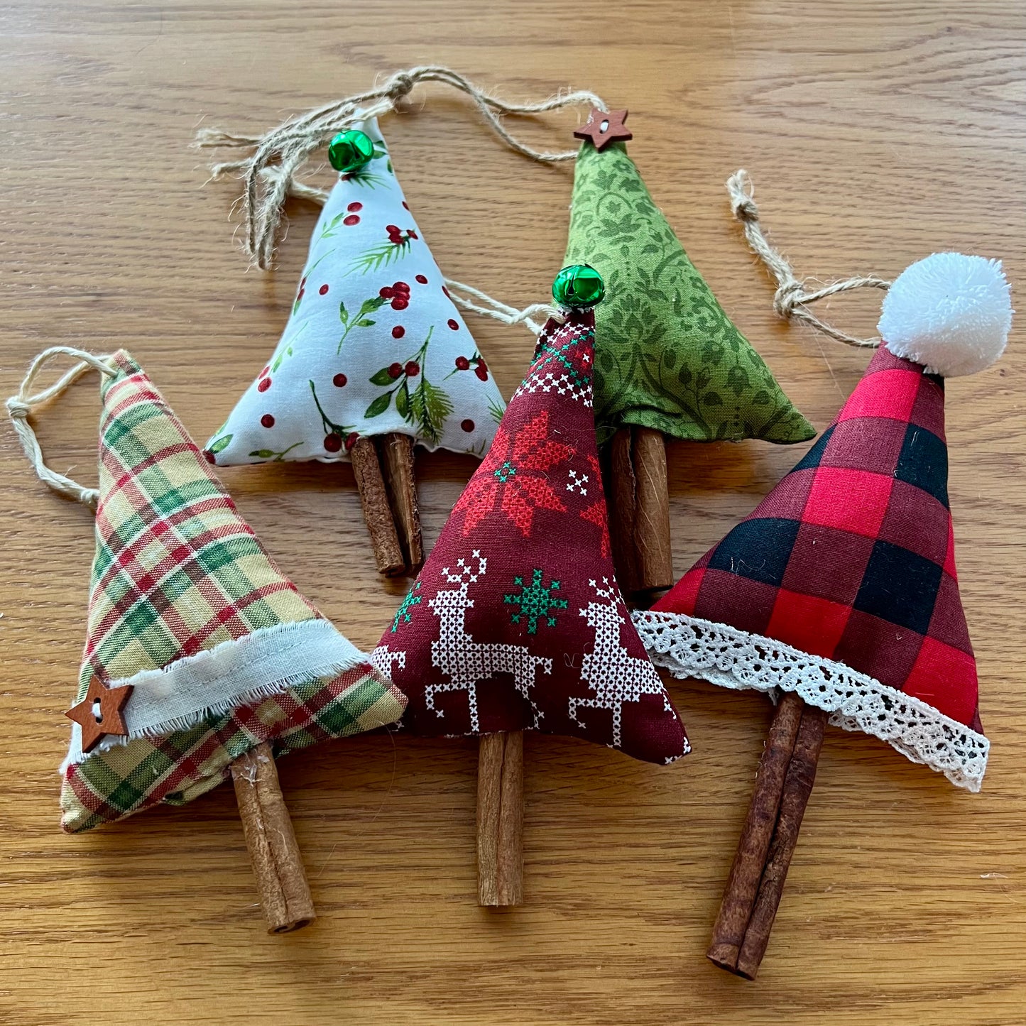Eclectic Spruce's handmade cinnamon stick trees are made with cotton fabric, stuffed with fiberfill, and embellished with real cinnamon sticks along with lace, buttons, bells, and other trim.  Sold mystery style, you will receive a curated collection of 5 unique trees picked out just for  you. This photo shows 5 trees arranged on a wooden surface.