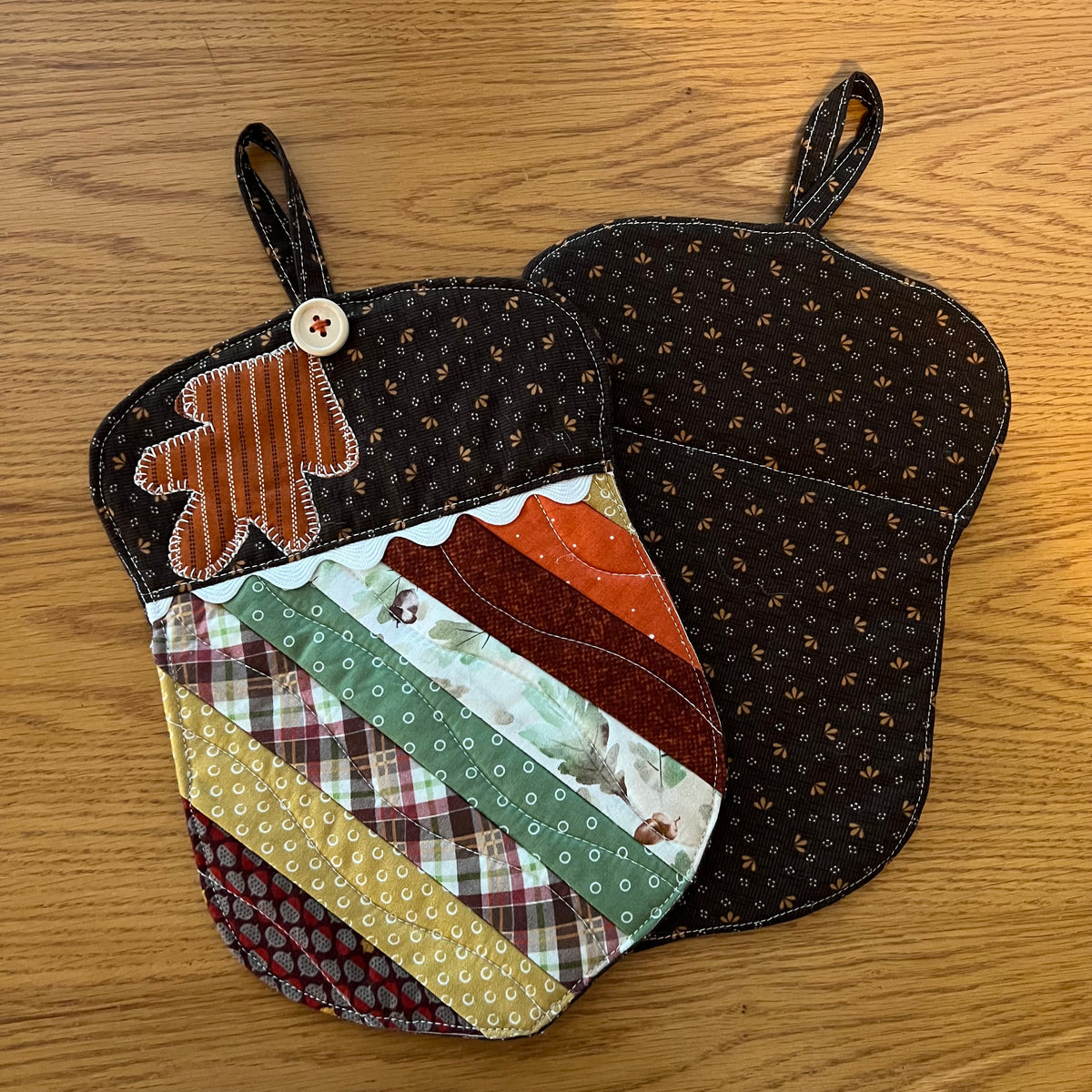 Two Eclectic Spruce acorn-shaped quilted potholder with hanging loops displayed on a wooden surface. One potholder is shown from the front, featuring autumn-themed patchwork fabrics, applique leaf design, wood button, and rick rack detailing. The other one is shown from the back side, featuring a dark brown small-scale floral cotton print.