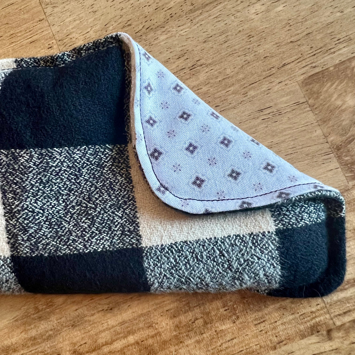 A close-up of the Nordic Check Weighted Eye Pillow by Eclectic Spruce. a flannel fabric with a black, white, and grey buffalo plaid pattern. The corner of the pillow is folded over, revealing a white cotton fabric with a small geometric pattern in black and grey on the reverse side.