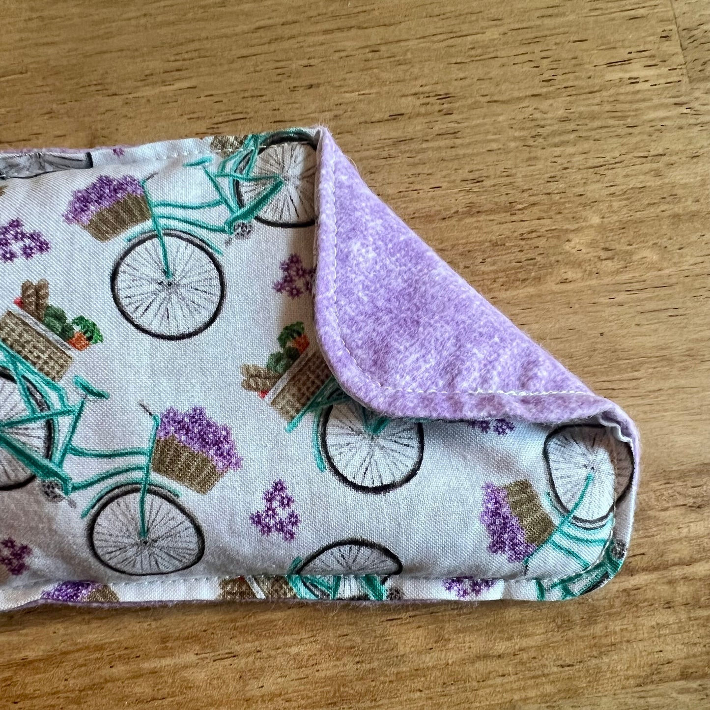 A close-up of a Petite Provence Weighted Eye Pillow by Eclectic Spruce. The front side is a white cotton fabric with teal bicycles, baskets of flowers, and purple blossoms. The corner of the pillow is folded over, revealing a light purple flannel fabric.