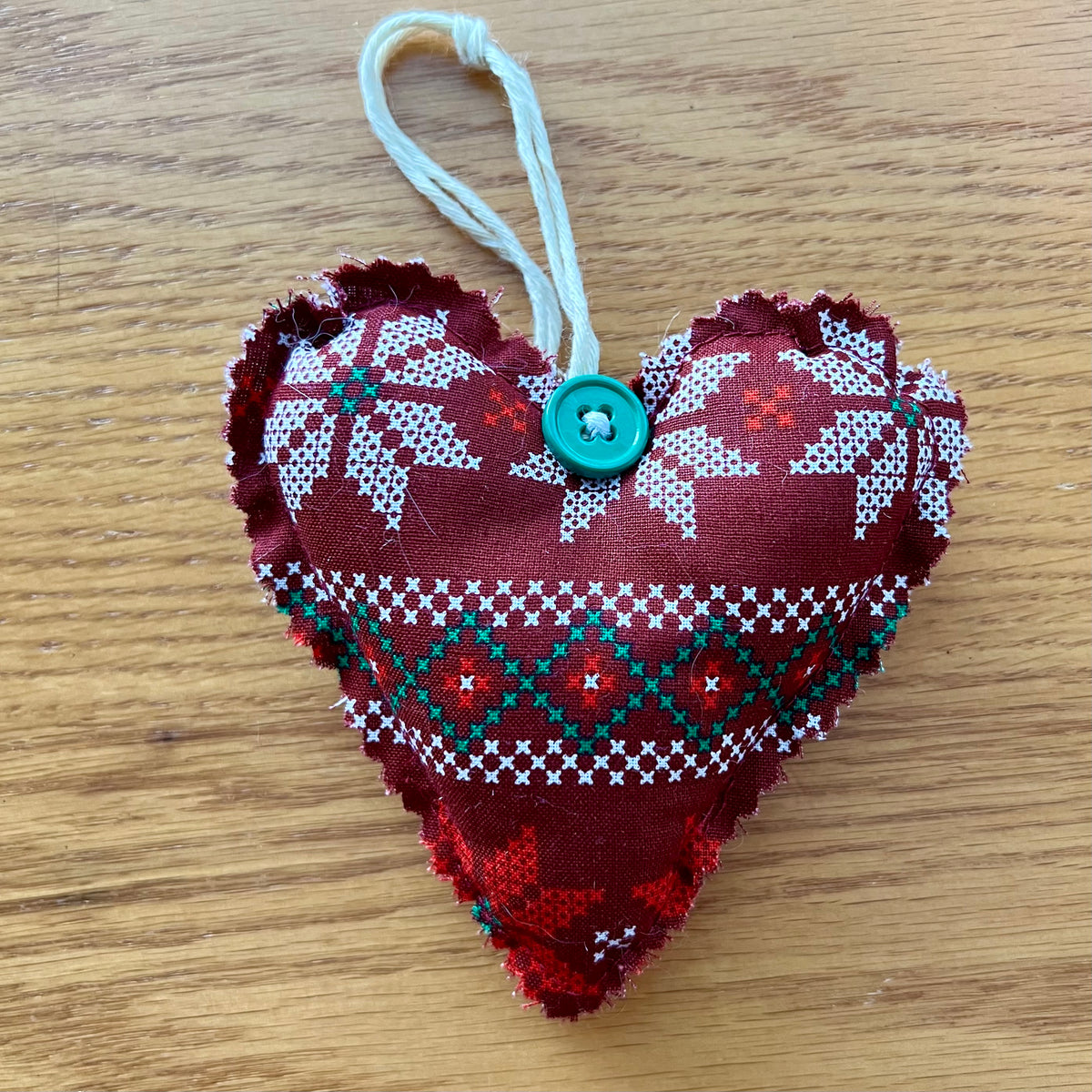 Eclectic Spruce's handmade fabric heart-shaped ornaments are made of cotton, stuffed with fiberfill, and embellished with lace, buttons, bells, and other trim.  Sold mystery style, you will receive a unique heart picked out just for  you. This photo shows ONE Eclectic Spruce heart ornament sitting on a wooden surface.