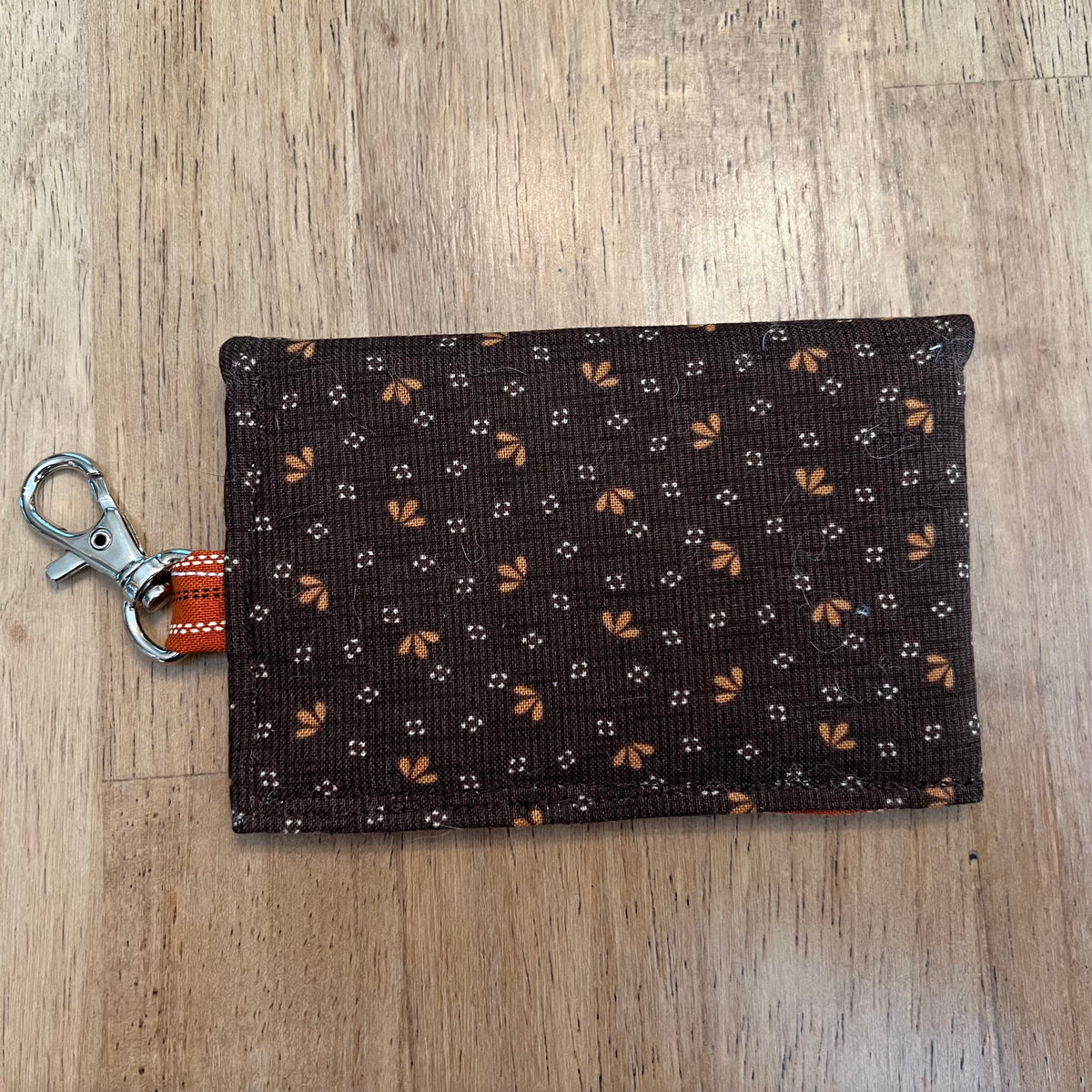 The back side of an Eclectic Spruce handcrafted mini snap wallet. It is made of a dark brown floral fabric, featuring a rust striped loop, and a silver metal swivel clasp.