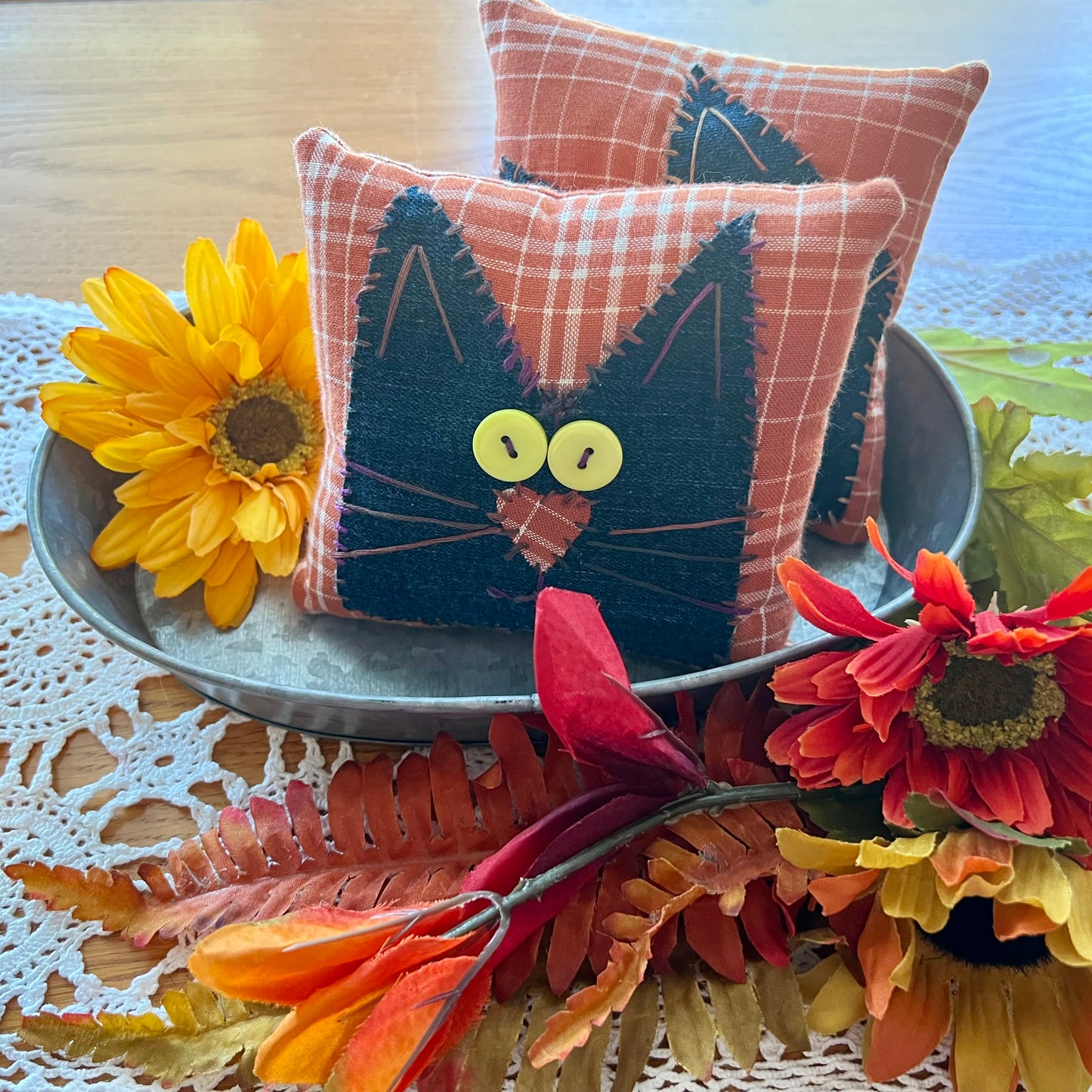 Two handmade Eclectic Spruce Purrfect Patchwork Cat pillows styled in an autumn setting. The front pillow features a black cat face applique on an orange and white plaid fabric background, complete with yellow button eyes and a stitched applique nose.