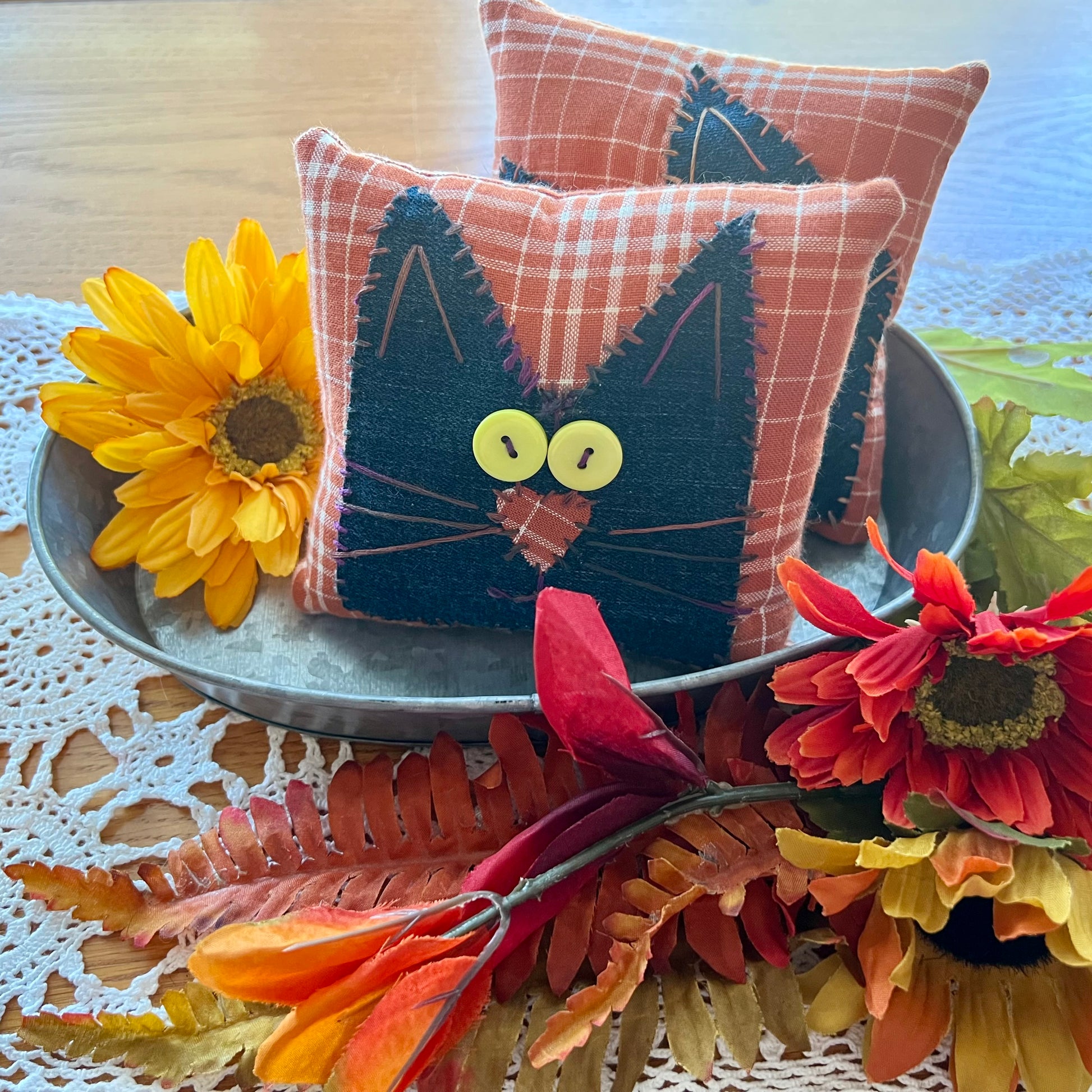 Two handmade Eclectic Spruce Purrfect Patchwork Cat pillows styled in an autumn setting. The front pillow features a black cat face applique on an orange and white plaid fabric background, complete with yellow button eyes and a stitched applique nose.