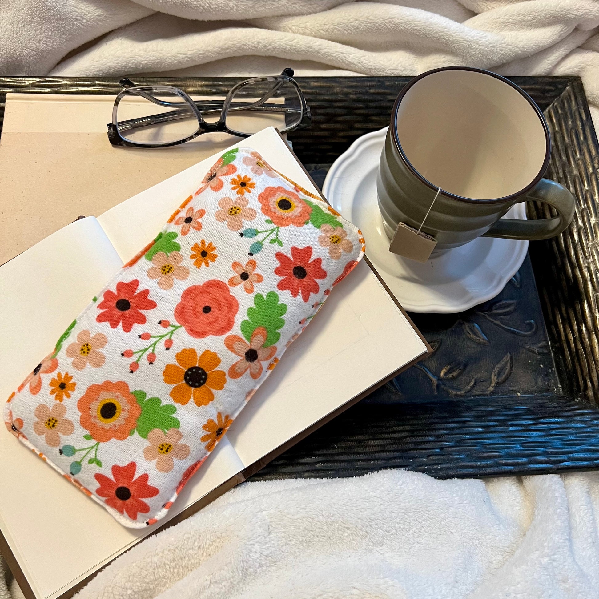 An Eclectic Spruce Retro Bloom Weighted Eye Pillow sitting on a tray with a book, mug, pair of glasses, and a cozy white blanket.