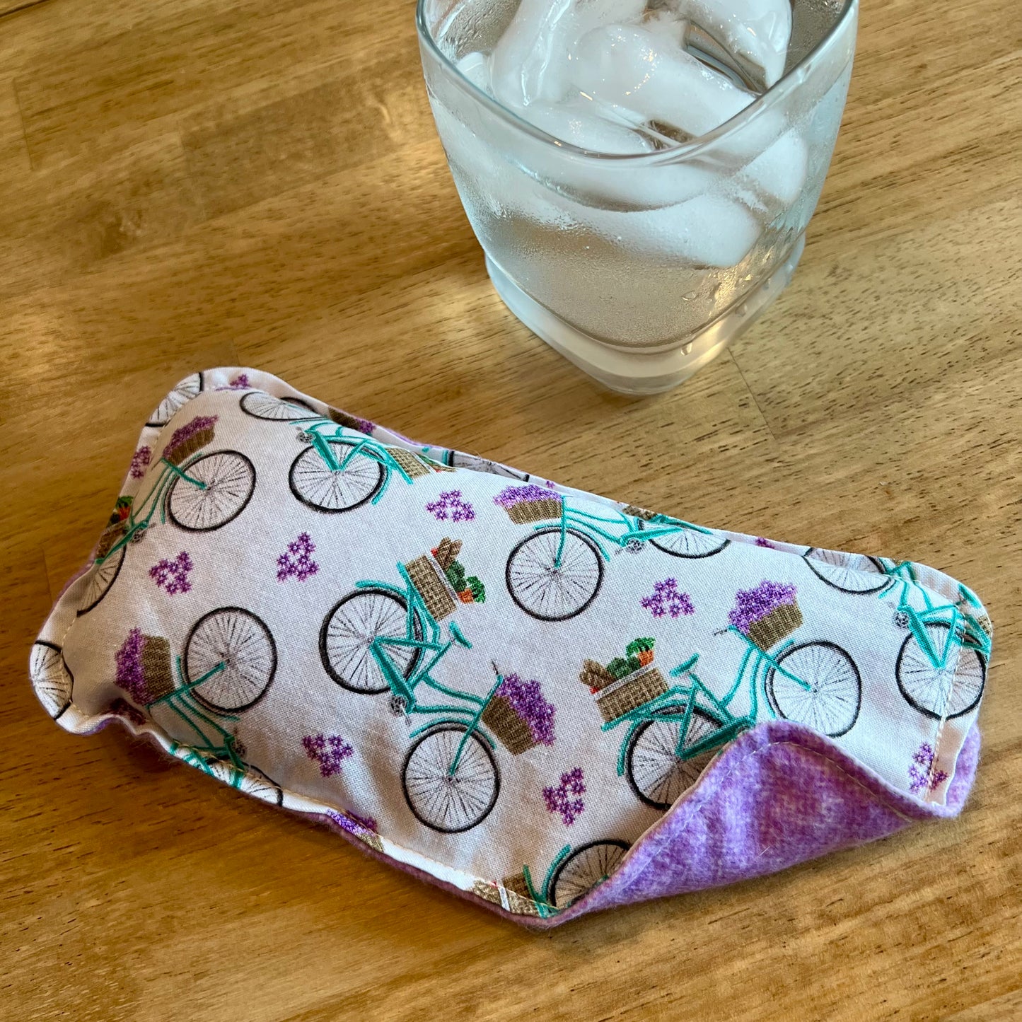 An Eclectic Spruce Petite Provence Weighted Eye pillow on a wood surface next to a glass of ice water.