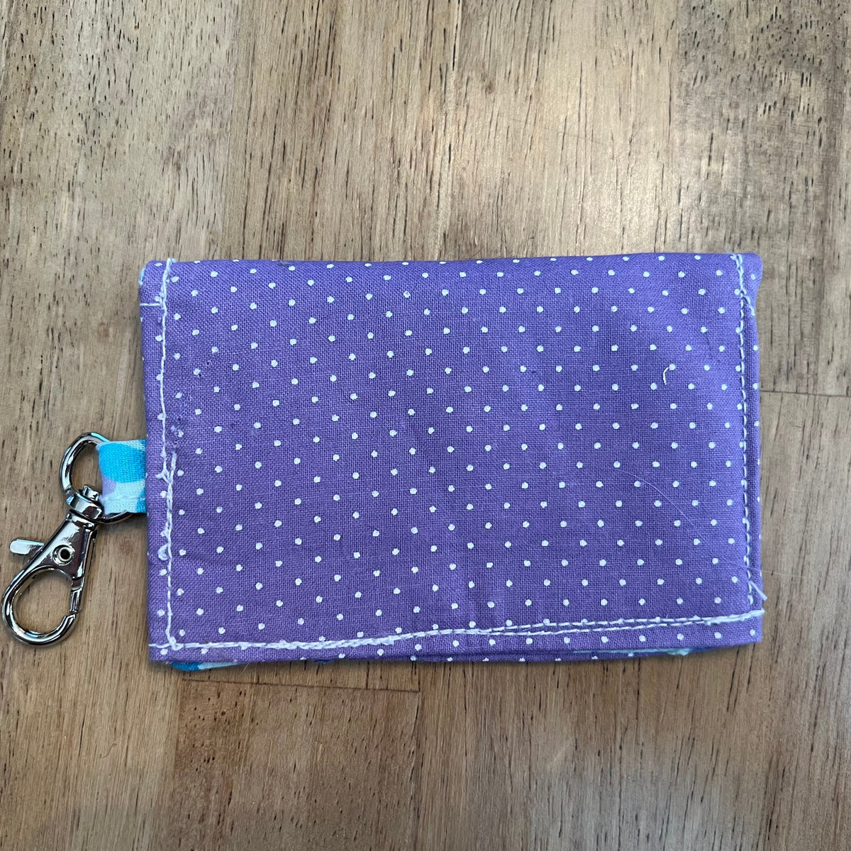 The back side of an Eclectic Spruce handcrafted mini snap wallet. It is made of purple fabric with white polka dots, featuring a blue floral fabric loop, and a silver metal swivel clasp.