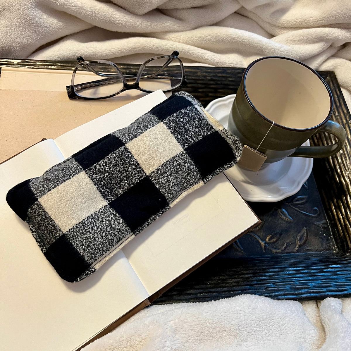 A Nordic Check Weighted Eye Pillow by Eclectic Spruce sitting on a tray with a book, mug, pair of glasses, and a cozy white blanket.
