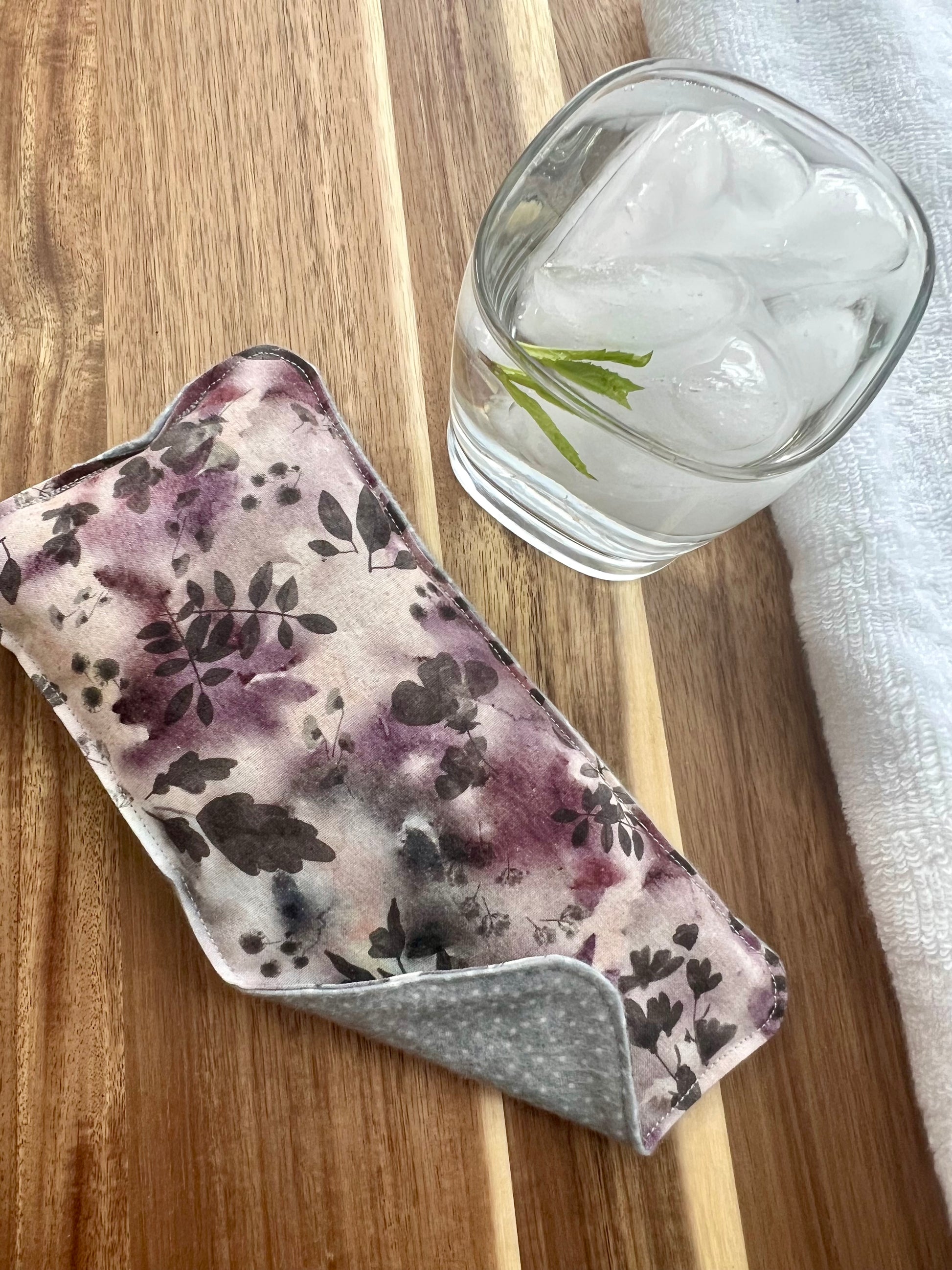 Weighted Eye Pillow on a wood surface next to a white towel and glass of ice water.