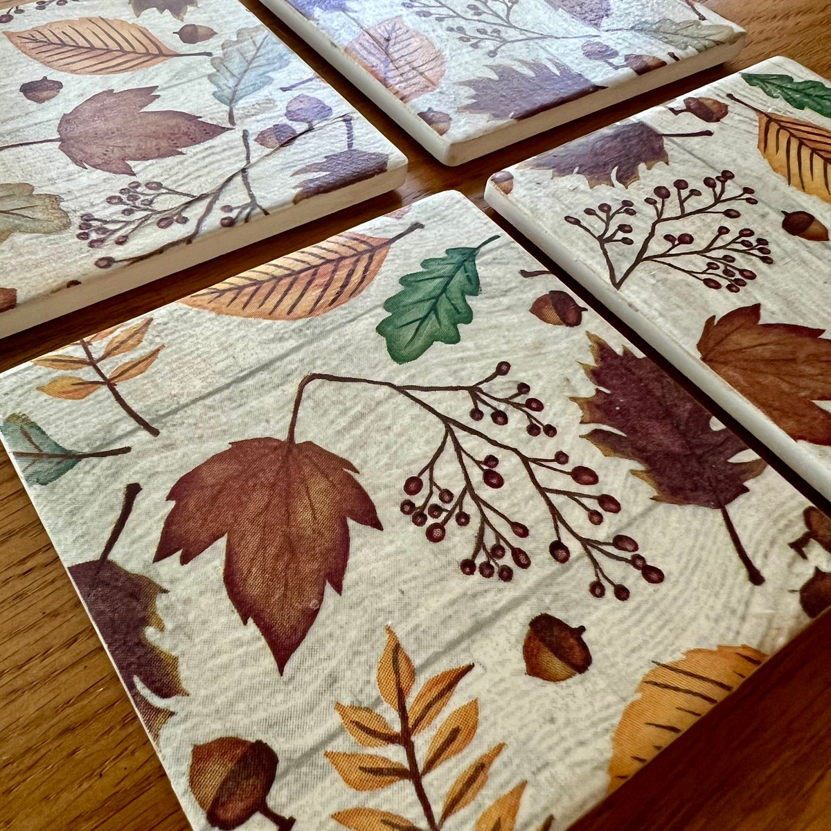 Four Eclectic Spruce square ceramic coasters with an autumn leaf pattern, arranged on a wooden table.