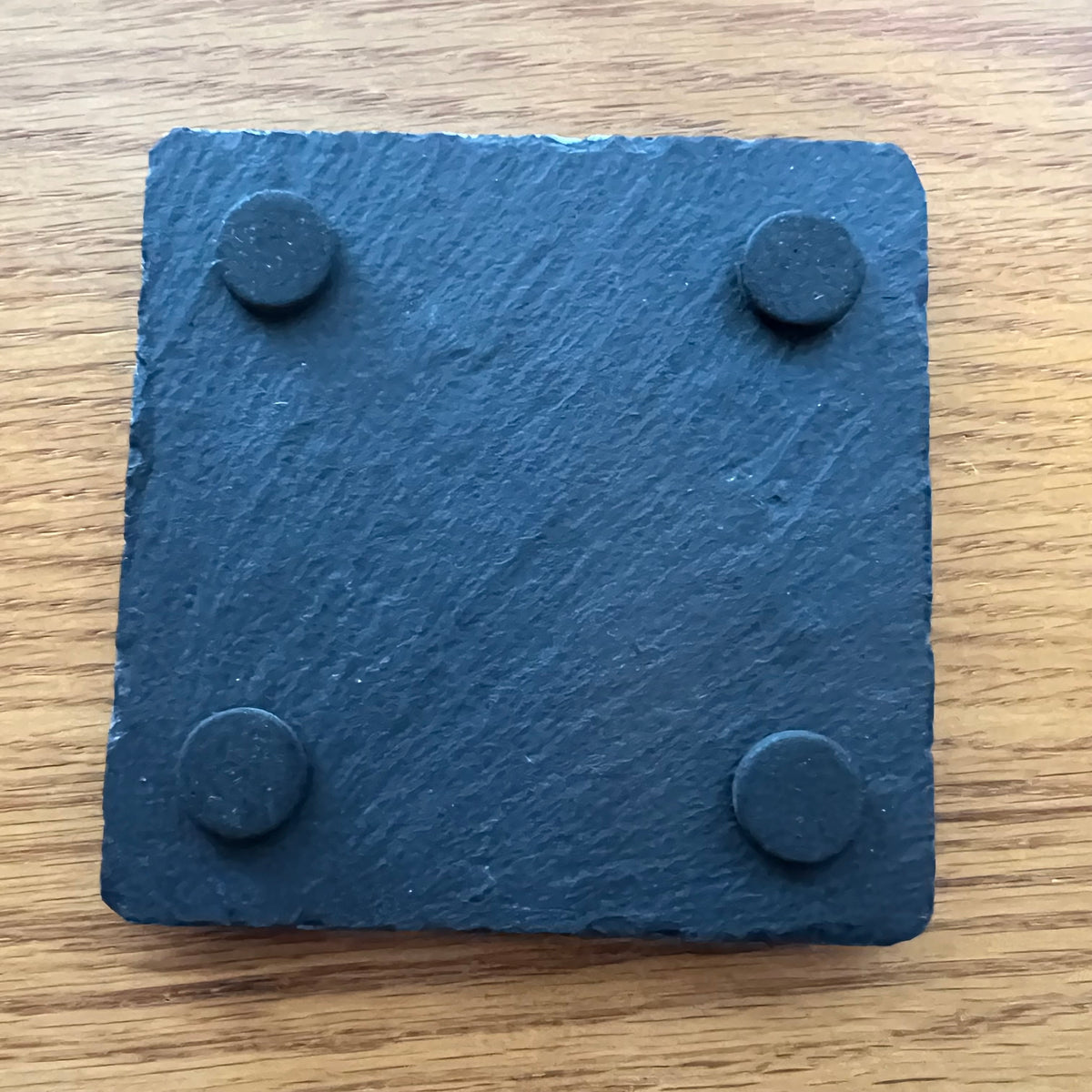 An Eclectic Spruce square black slate coaster with four circular rubber feet on the underside, shown on a wooden surface.