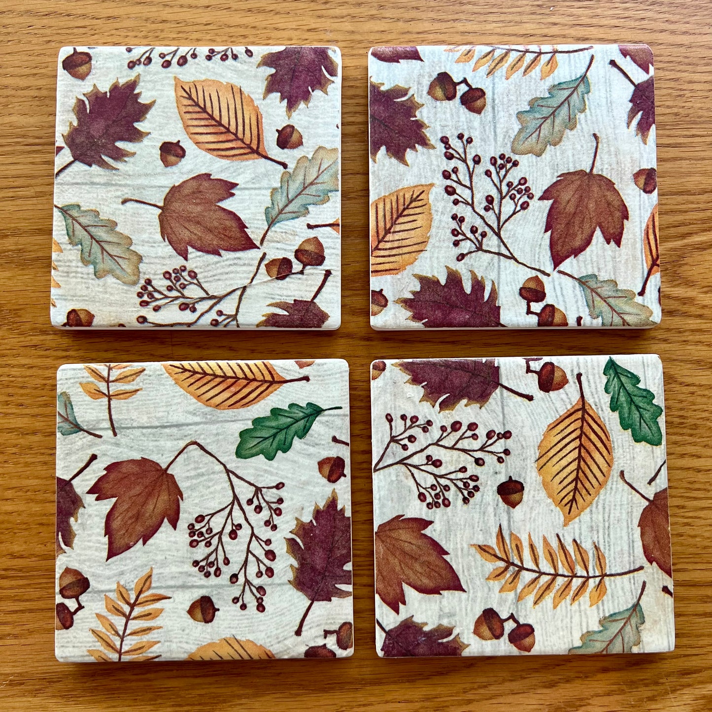 Four Eclectic Spruce ceramic square coasters with decoupaged with an autumn leaf pattern, arranged in a 2x2 grid on a wooden tabletop.