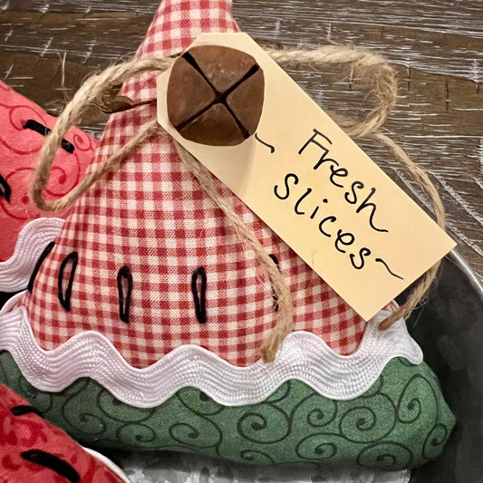 Close-up of a fabric watermelon slice with a 'Fresh slices' tag, rusty bell, and twine bow.