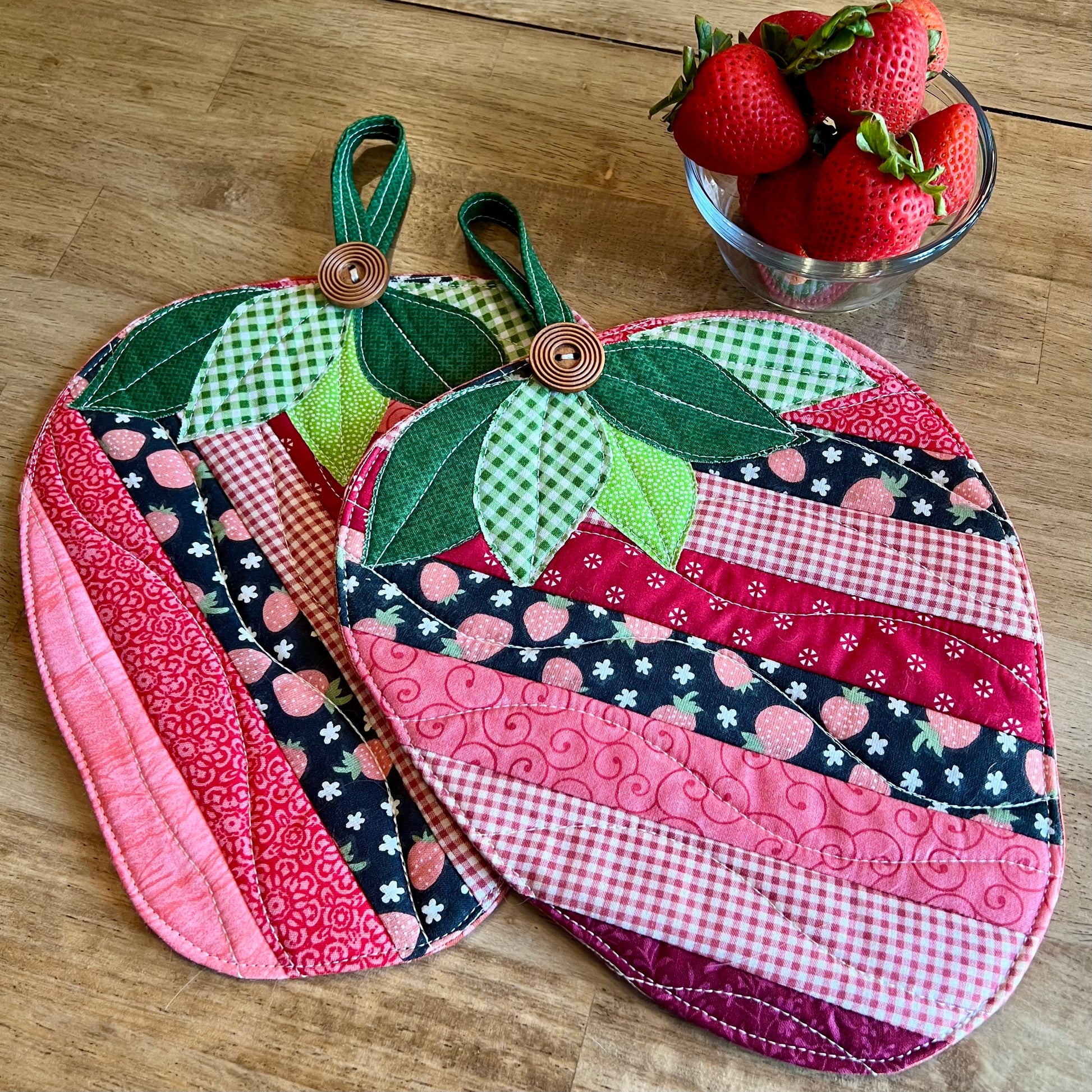 Two Eclectic Spruce Sweet Strawberry hot pads (strawberry shaped, quilted potholders with green leaf tops and wooden button handles), displayed on a wooden tabletop next to a glass bowl of fresh strawberries.
