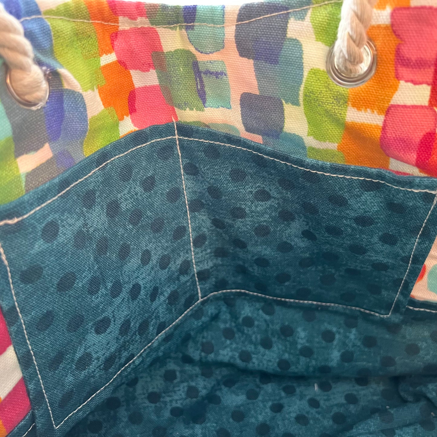 Close-up of the Eclectic Spruce rope-handled tote bag showing a colorful patchwork pattern on the upper section, two dark teal polka-dot inner pockets, and white rope handles with metal grommets.