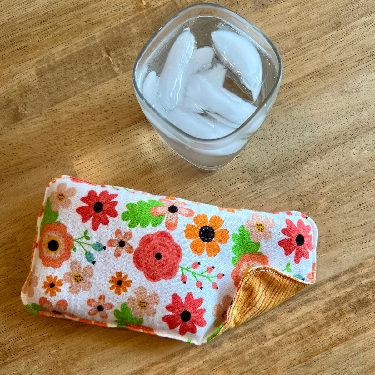 An Eclectic Spruce Retro Bloom Weighted Eye pillow on a wood surface next to a glass of ice water.