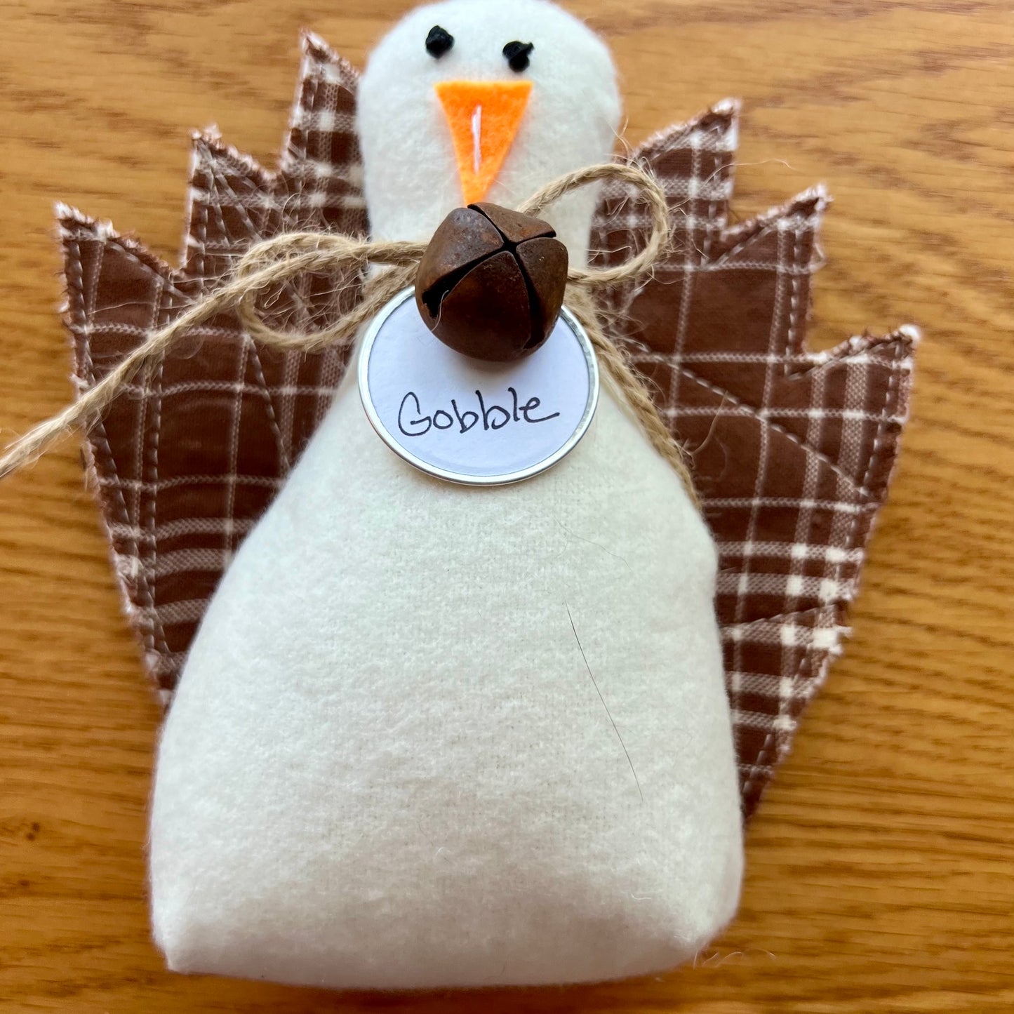 An Eclectic Spruce handmade fabric Thanksgiving turkey sitting on a wooden table. The turkey features a white flannel body, textile tail feathers, an orange felt beak, black stitched eyes, a brown rusty bell on a twine bow, and a round white tag that reads "Gobble."