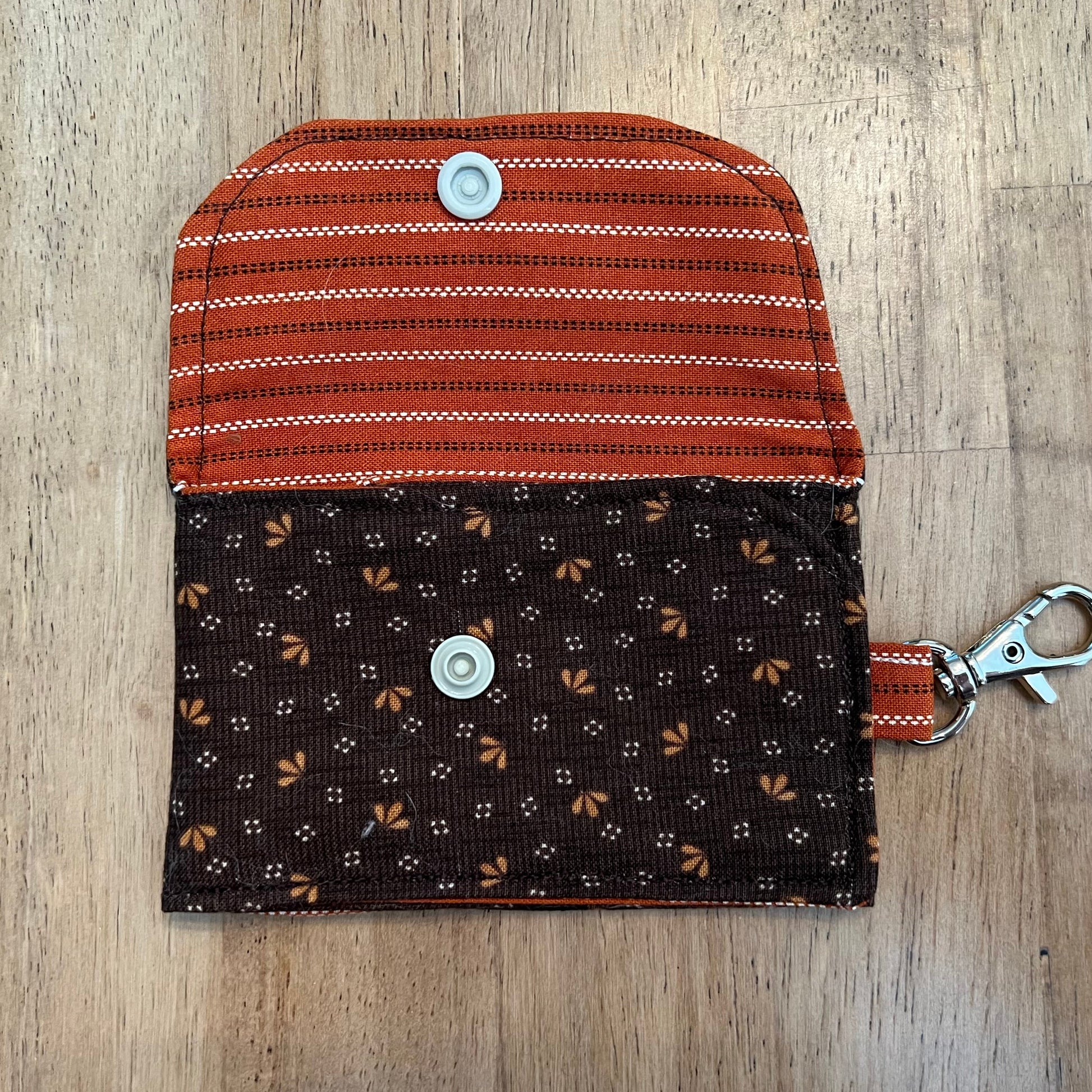 Eclectic Spruce handcrafted mini card holder shown in an open position with a dark brown floral exterior and a rust striped interior. The wallet contains a beige snap closure and a silver metal swivel clasp for convenience and security.