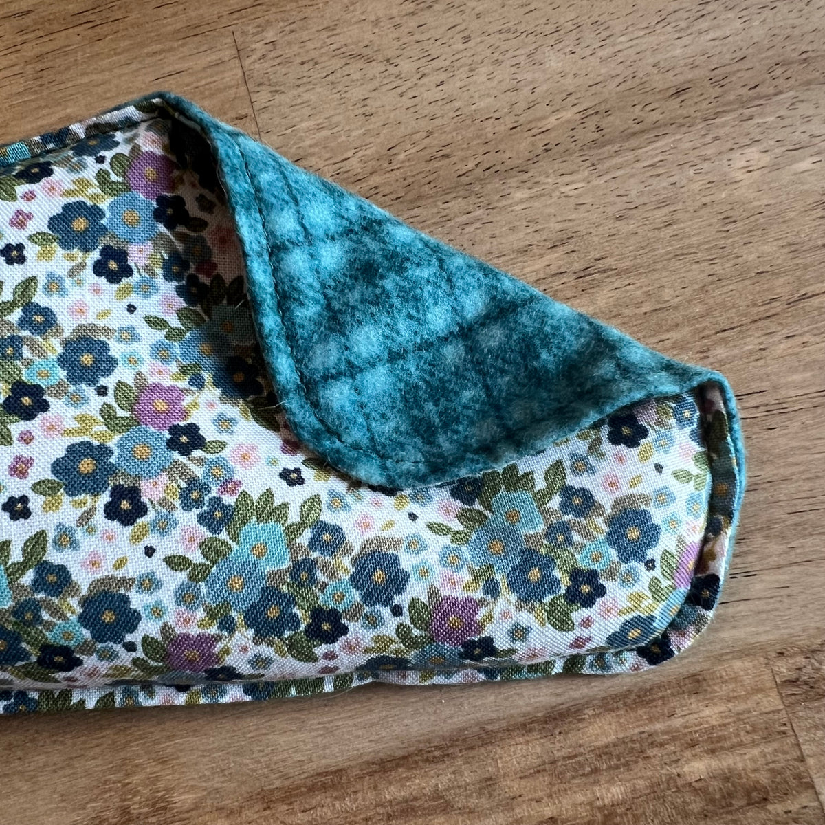 A close-up of the Wildflower Glen Weighted Eye Pillow by Eclectic Spruce. This pillow features a white cotton fabric with a colorful floral print in blue, purple, and green. A corner of the pillow is folded over, revealing a plaid flannel in shades of teal.