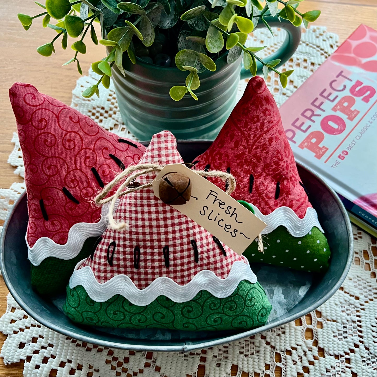 Three decorative fabric watermelon slices on a metal tray with a plant and book in the background.