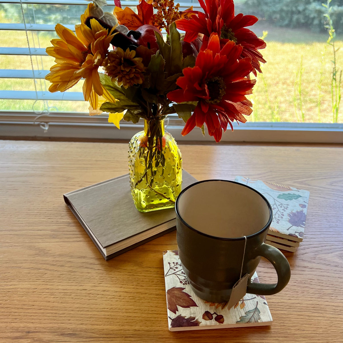 A lifestyle shot showing four Eclectic Spruce ceramic coasters in action. The coasters are square with a cozy autumn leaves print. A dark gray ceramic mug with a tea bag hanging from the handle sits on one coaster on a wooden table. The other three coasters are stacked neatly behind the mug. Nearby is a yellow textured glass vase filled with red, orange, and yellow flowers resting on a gray book, with a window and outdoor grass and spruce trees visible in the background.