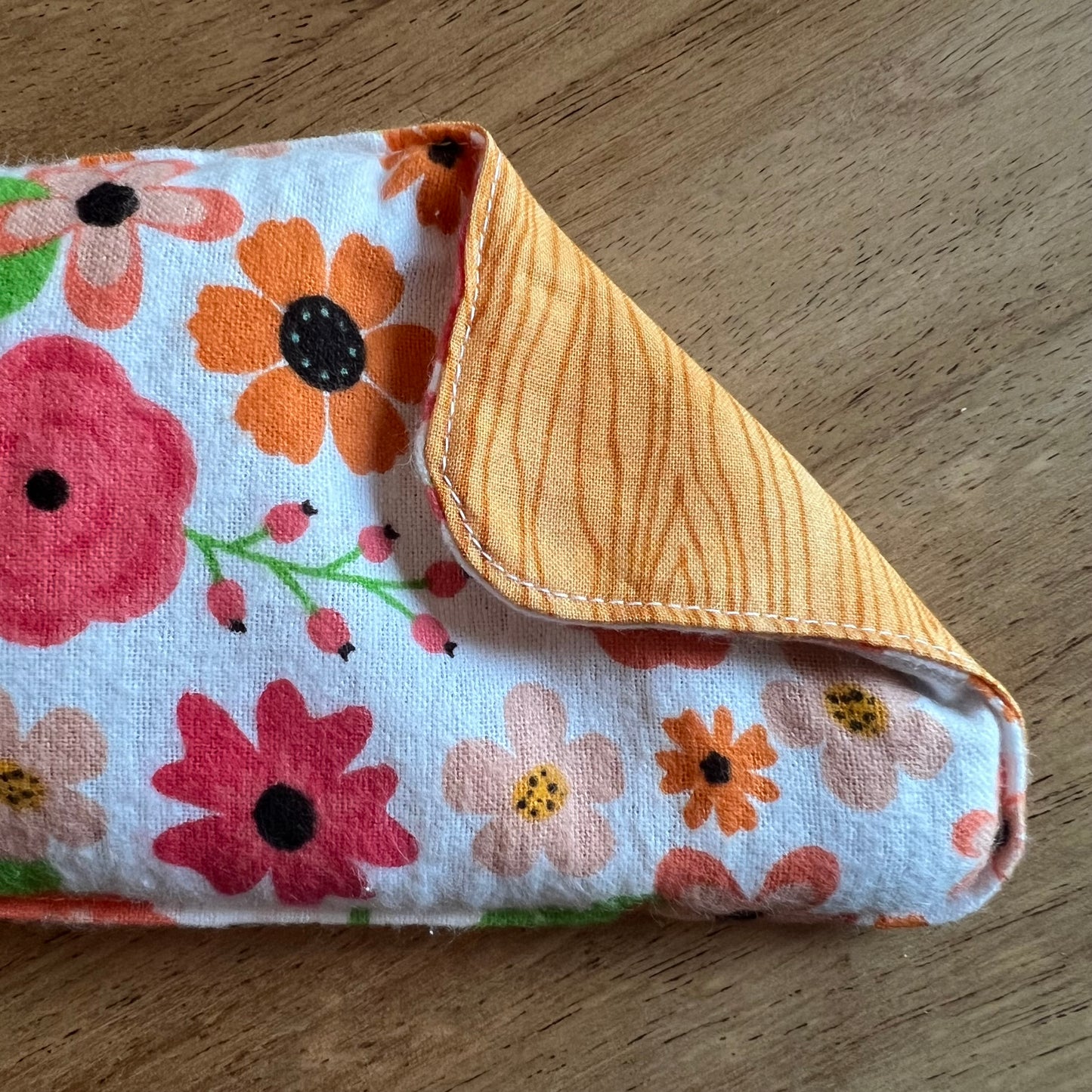 A close-up of the Retro Bloom Weighted Eye Pillow by Eclectic Spruce. The front side is a white flannel with colorful floral print. The corner of the pillow is folded over, revealing an orange-brown wood grain patterned cotton fabric on the reverse side.
