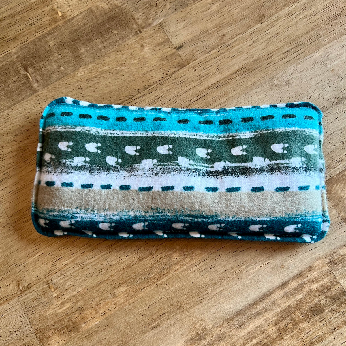 Dual-sided Alpine Path Weighted Eye Pillow by Eclectic Spruce. This pillow  features a striped pattern with animal tracks in turquoise, dark green, white, and beige.
