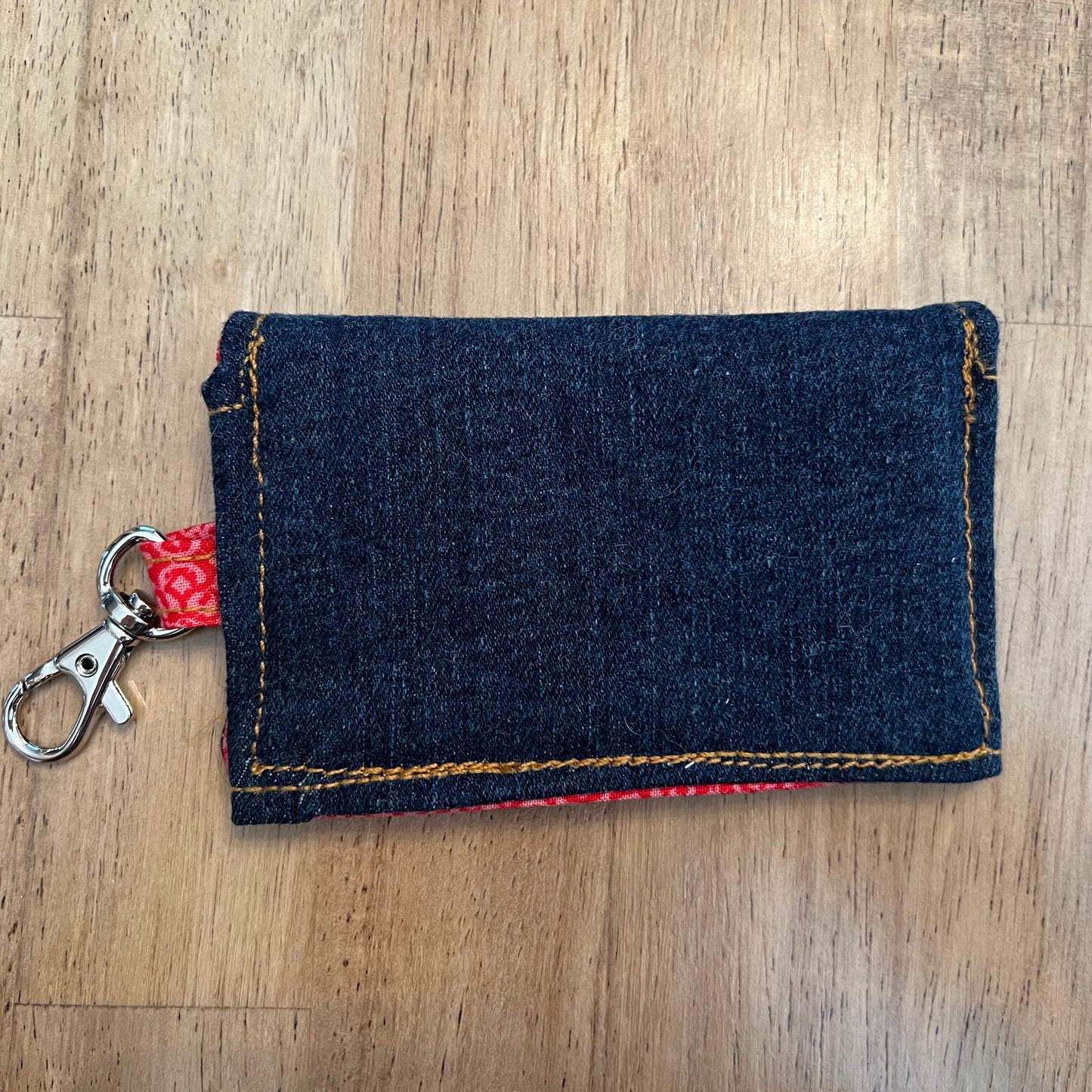 The back side of an Eclectic Spruce handcrafted mini snap wallet. It is made of dark blue denim, featuring an orange contrasting stitching, a red floral fabric loop, and a silver metal swivel clasp.