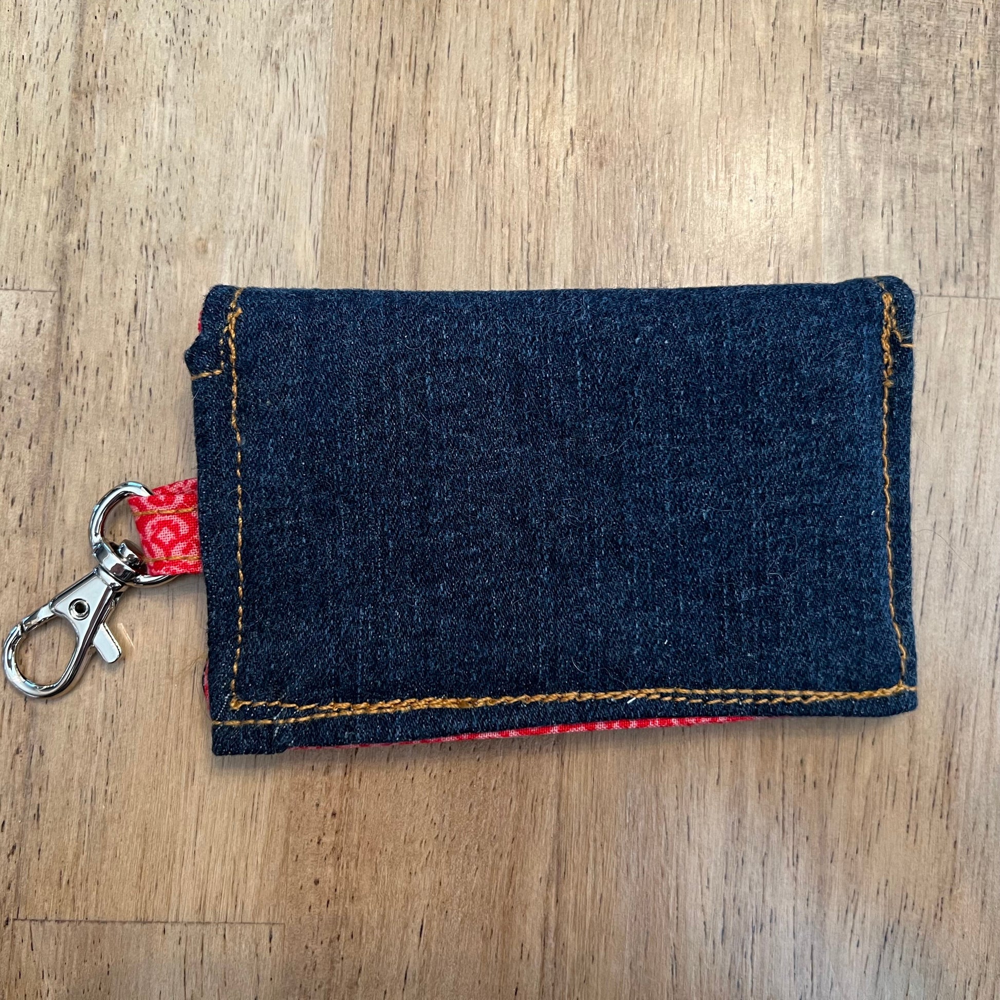 The back side of an Eclectic Spruce handcrafted mini snap wallet. It is made of dark blue denim, featuring an orange contrasting stitching, a red floral fabric loop, and a silver metal swivel clasp.