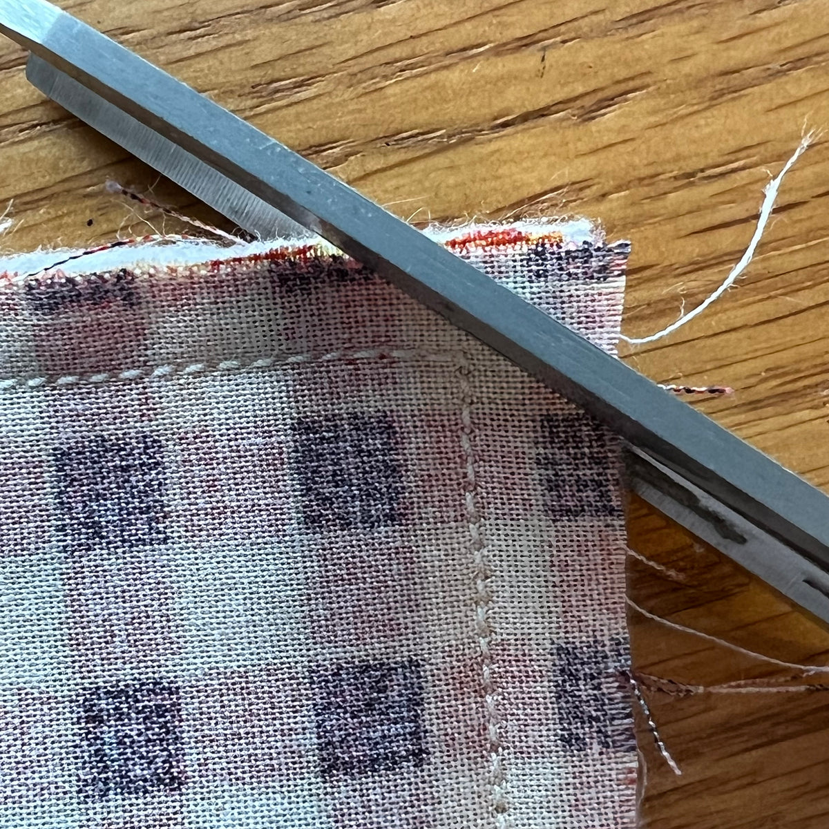 Close-up of checkered fabric being trimmed with a metal cutting tool on a wooden surface. This is an example photo from the Eclectic "Purrfect Patchwork Cat Pillows" sewing pattern.