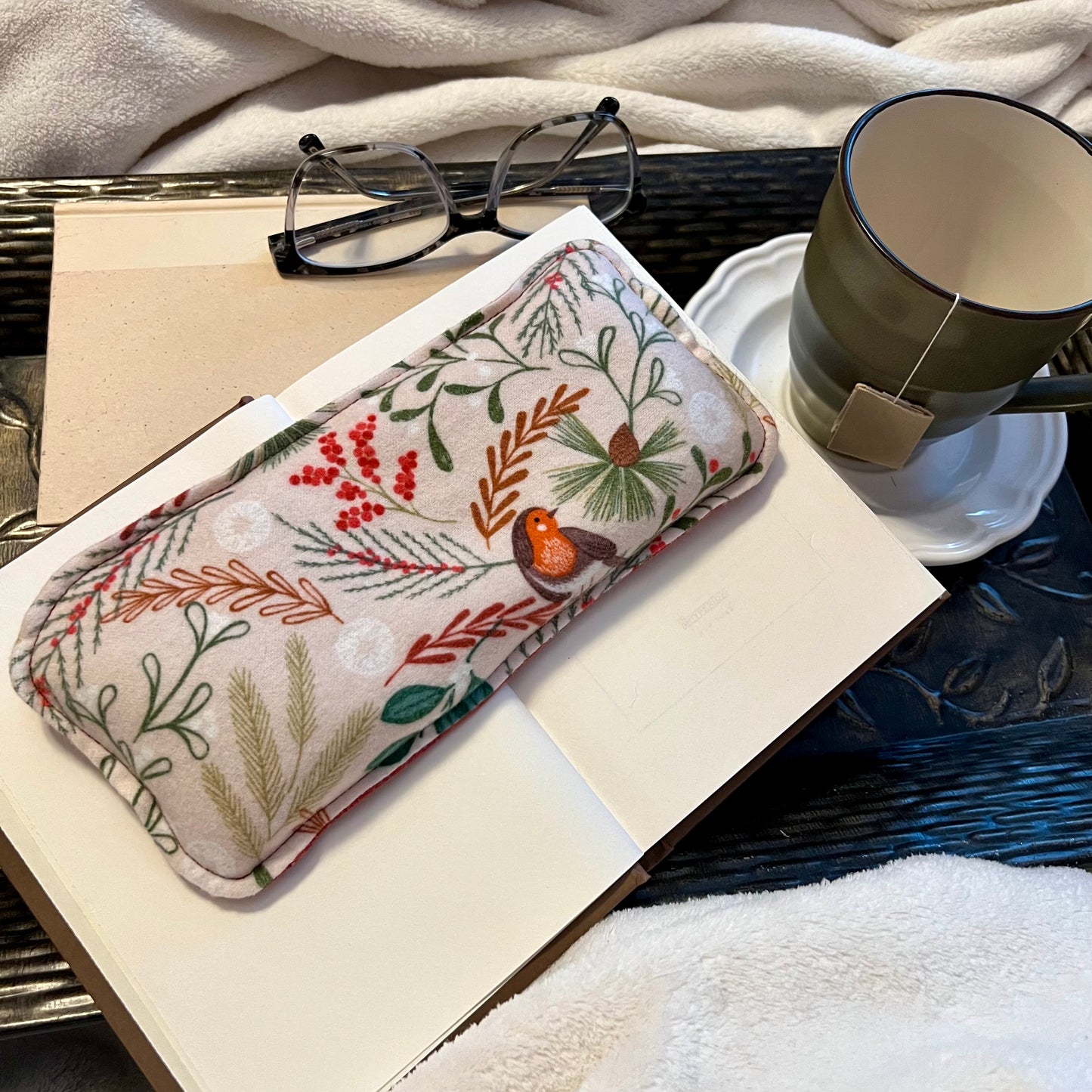 A Woodland Bough Eclectic Spruce Weighted Eye Pillow sitting on a tray with a book, mug, pair of glasses, and a cozy white blanket.