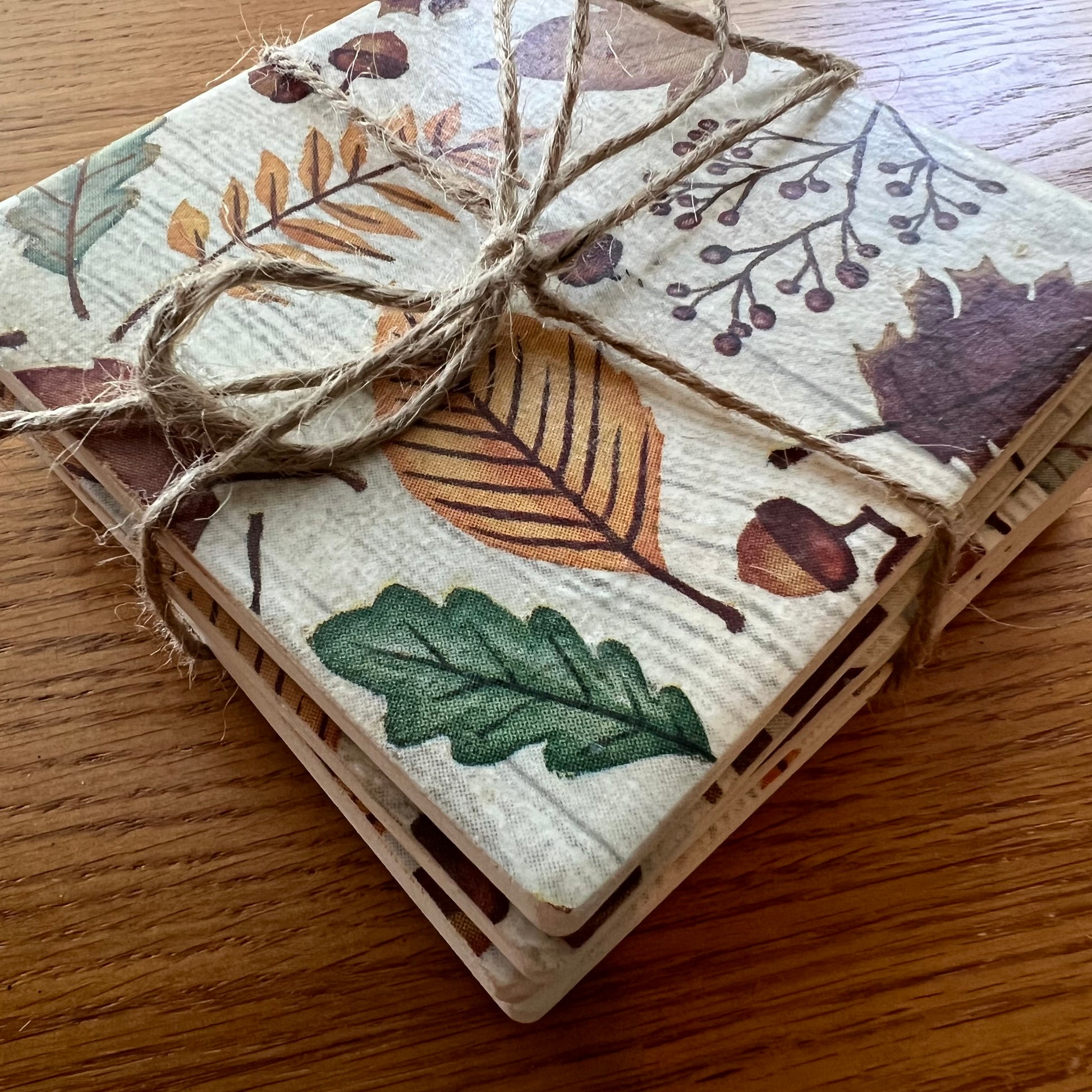 A stack of four Eclectic Spruce ceramic square coasters with a rustic autumn leaf pattern, tied together with natural jute twine, resting on a wooden table.