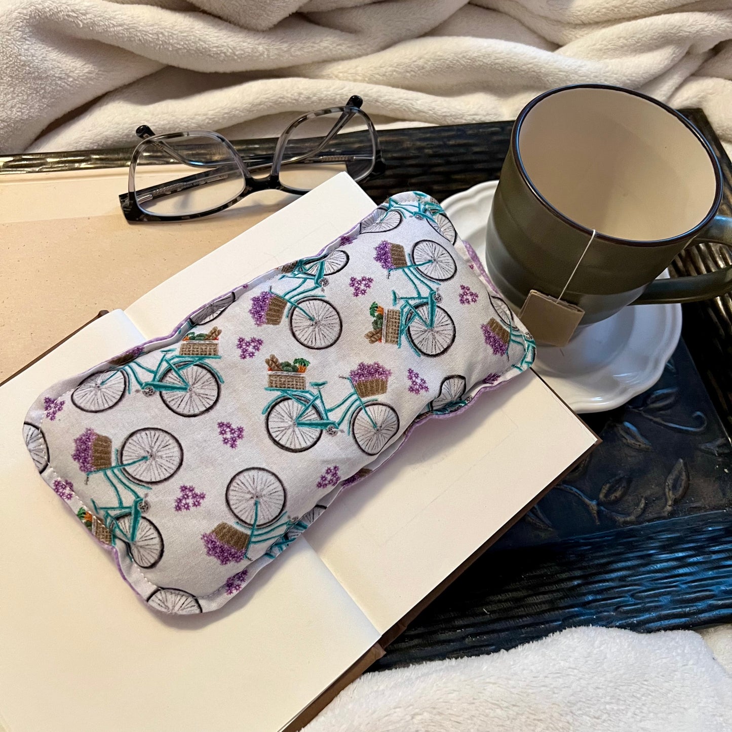 An Eclectic Spruce Petite Provence Weighted Eye Pillow sitting on a tray with a book, mug, pair of glasses, and a cozy white blanket.