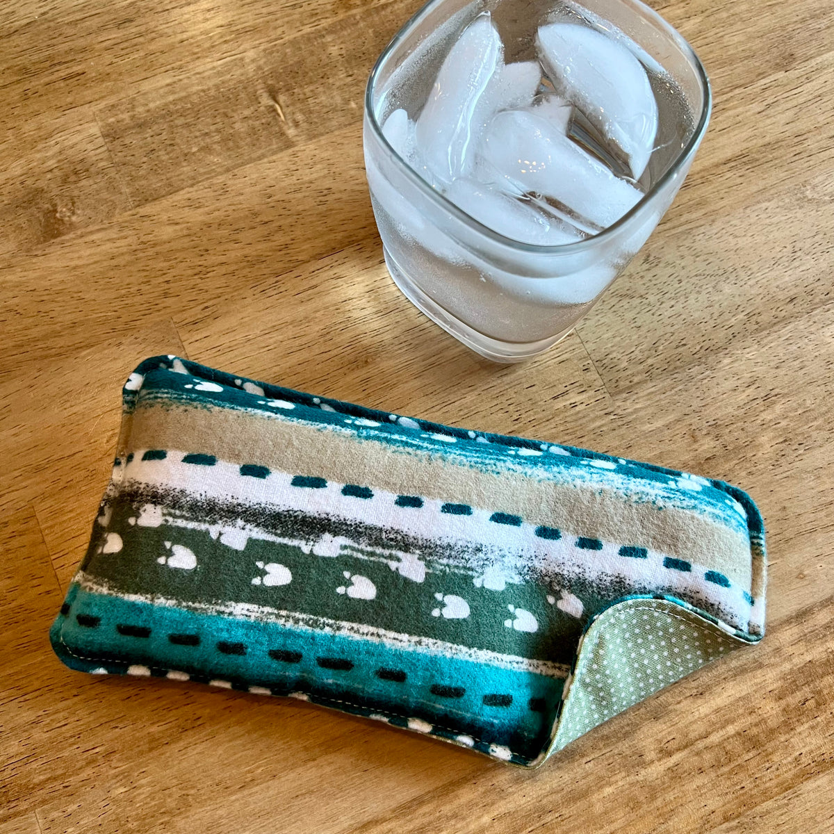 An Eclectic Spruce Alpine Path Weighted Eye pillow on a wood surface next to a glass of ice water.
