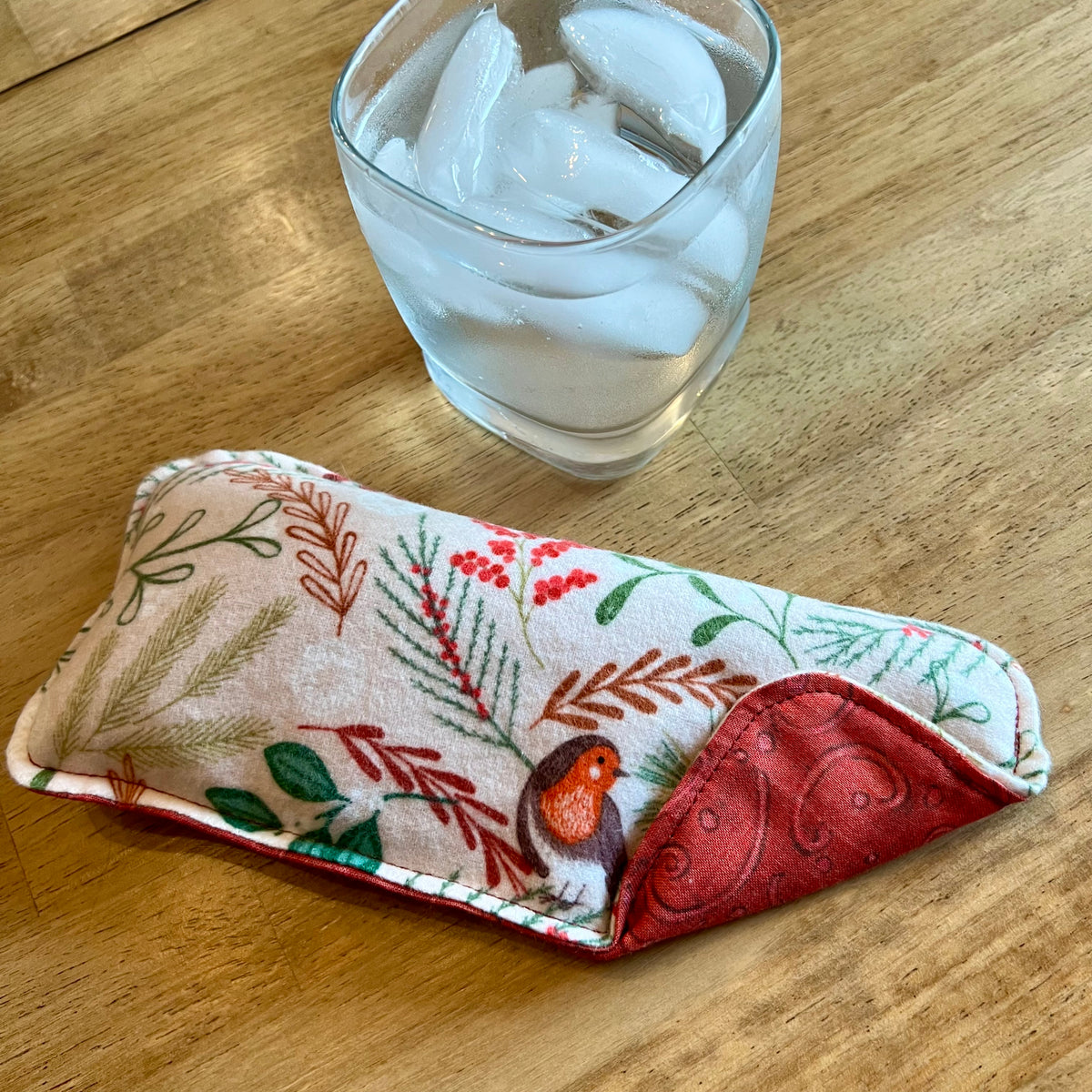 An Eclectic Spruce Woodland Bough Weighted Eye pillow on a wood surface next to a glass of ice water.
