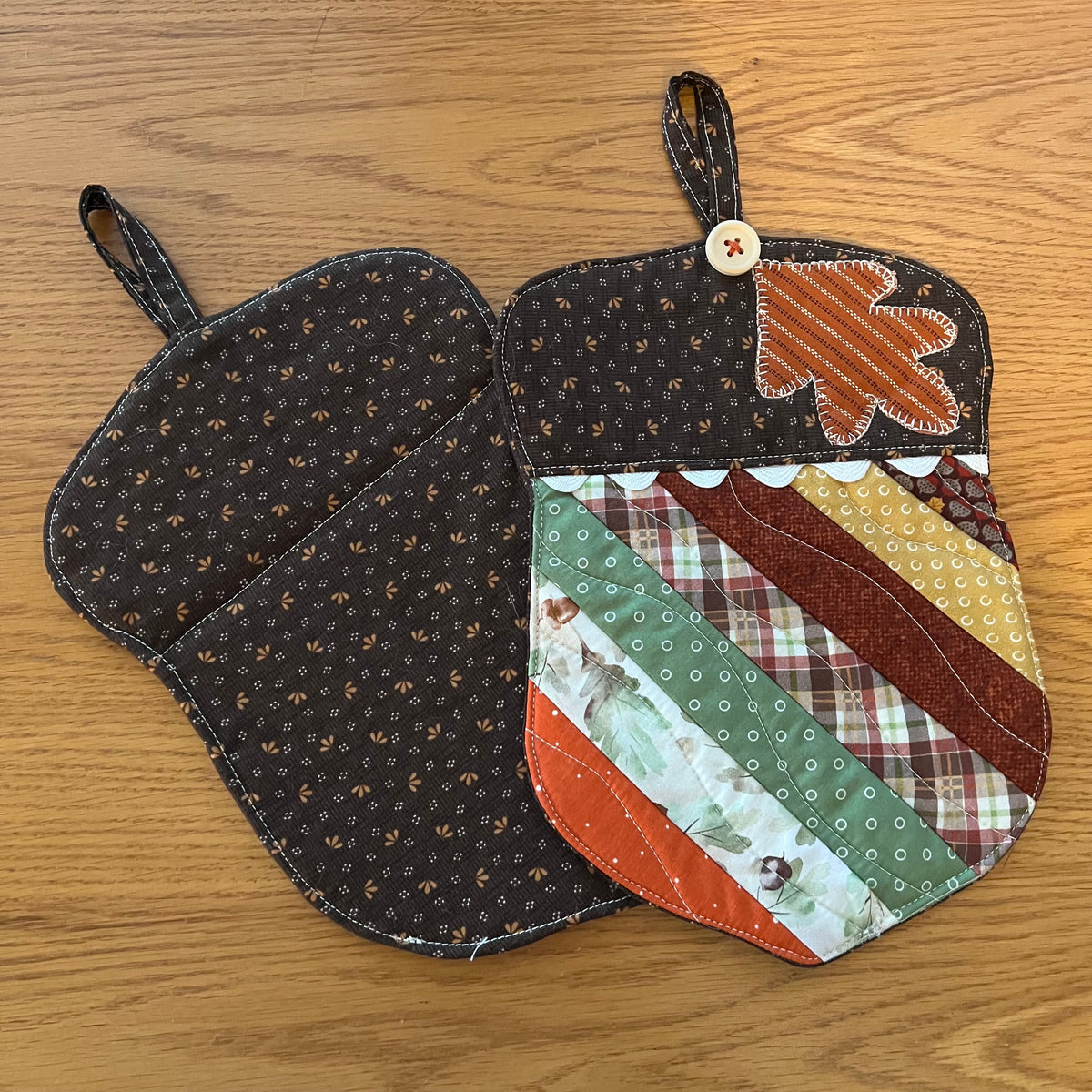 Two Eclectic Spruce acorn-shaped quilted potholder with hanging loops displayed on a wooden surface. One potholder is shown from the front, featuring autumn-themed patchwork fabrics, applique leaf design, wood button, and rick rack detailing. The other one is shown from the back side, featuring a dark brown small-scale floral cotton print.