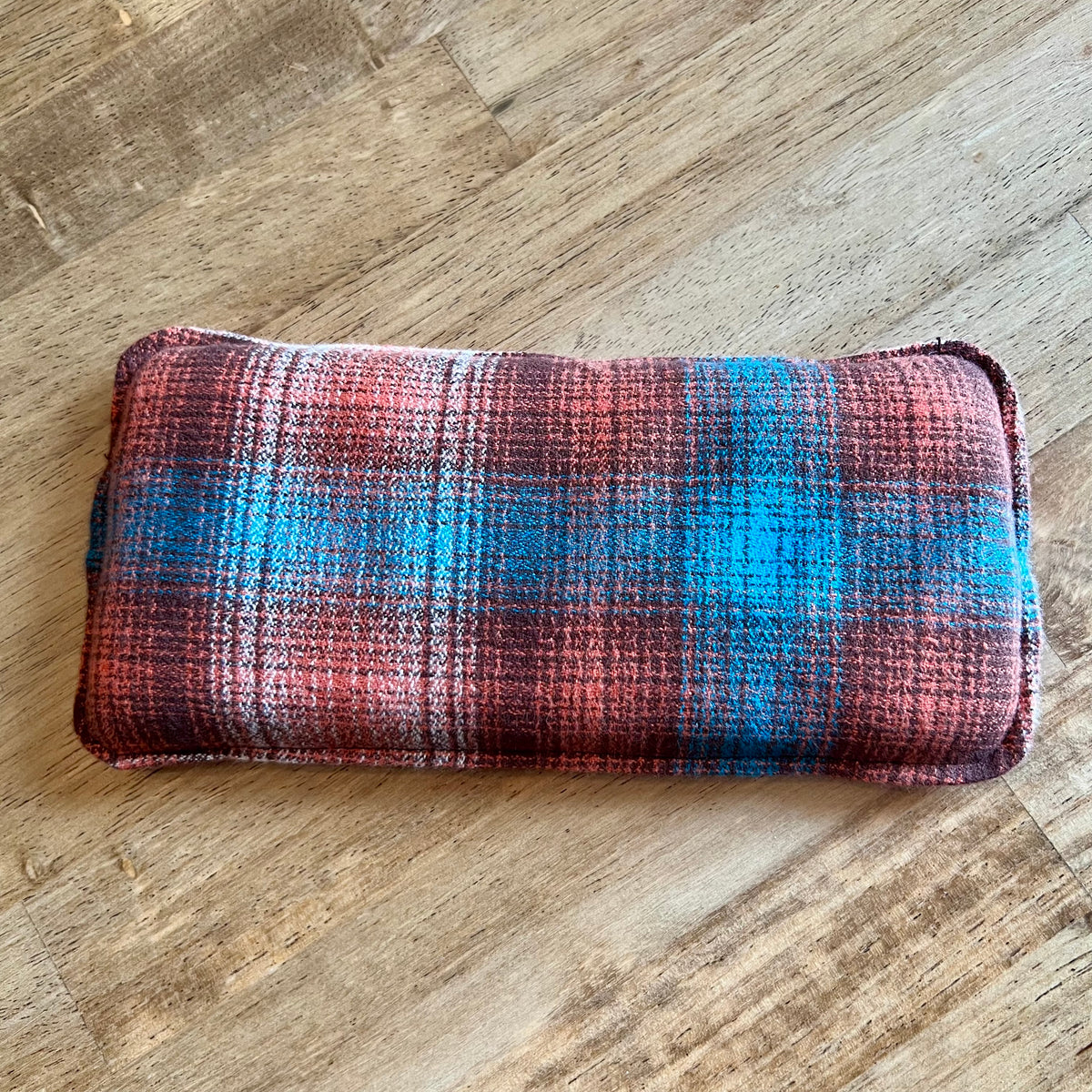 A dual-sided Copper Ridge Weighted Eye Pillow by Eclectic Spruce. This pillow features a plaid pattern in rust orange, teal, and dark brown.