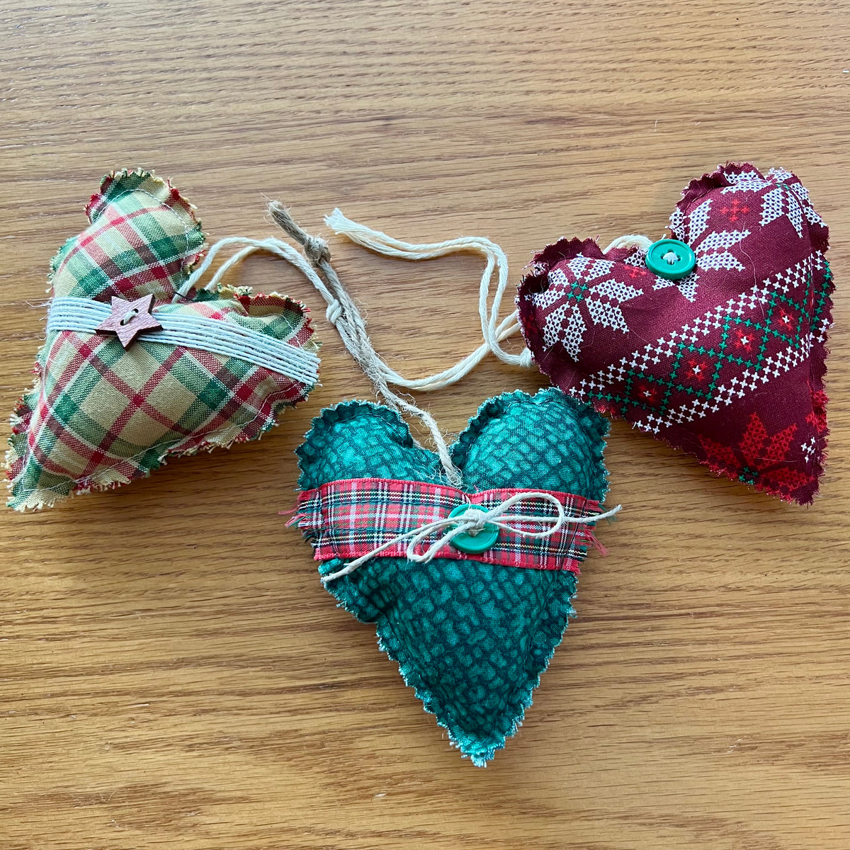 Eclectic Spruce's handmade fabric heart-shaped ornaments are made of cotton, stuffed with fiberfill, and embellished with lace, buttons, bells, and other trim.  Sold mystery style, you will receive a curated collection of 3 unique hearts picked out just for you. This photo shows 3 Eclectic Spruce heart ornaments sitting on a wooden surface.