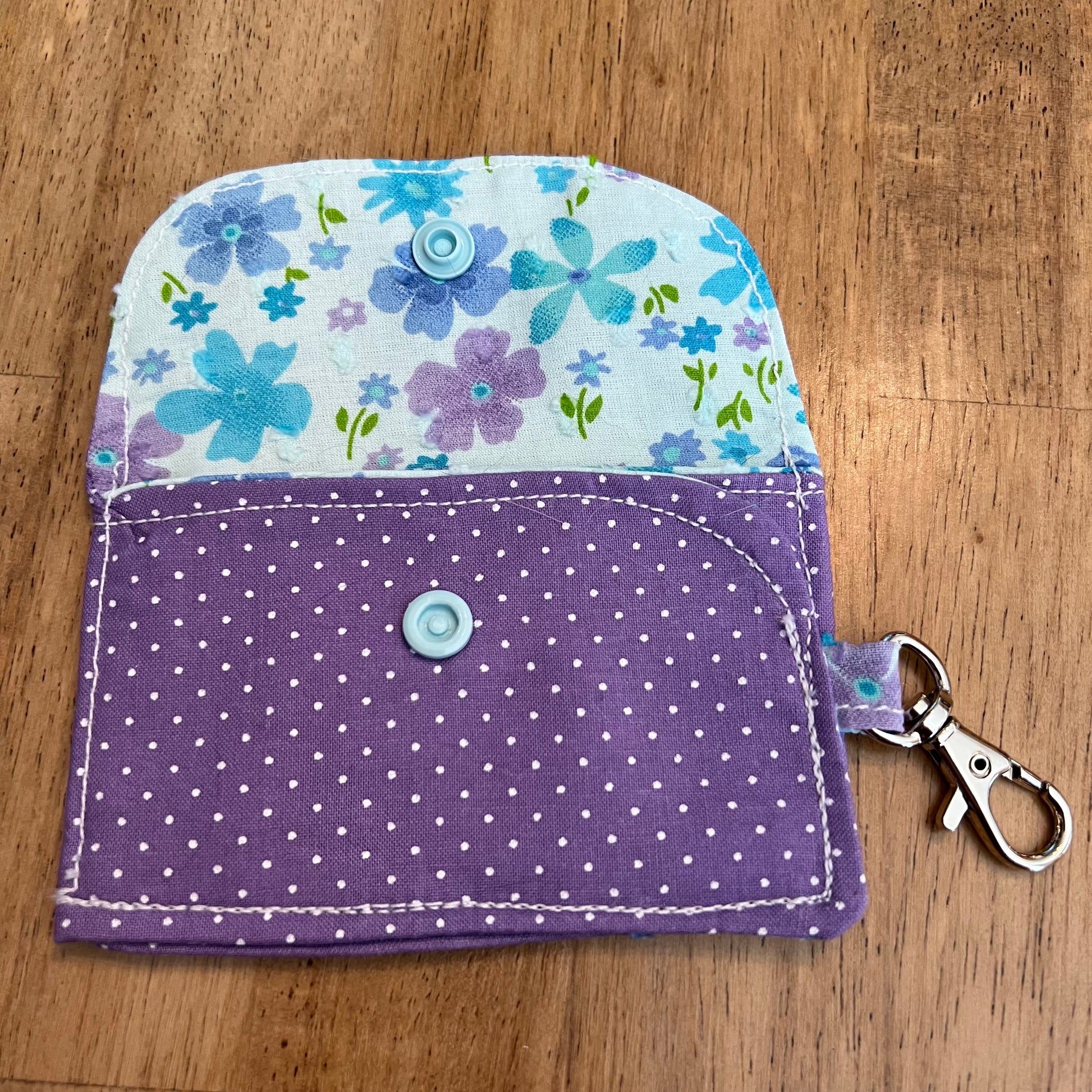 Eclectic Spruce handcrafted mini card holder wallet shown in an open position with a purple polka dot patterned exterior and a light blue floral patterned interior. The wallet contains a sky blue snap closure and a silver metal swivel clasp for convenience and security.