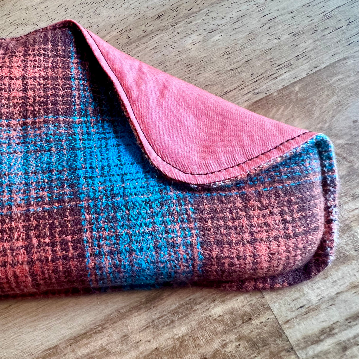 A close-up of the Copper Ridge Weighted Eye Pillow by Eclectic Spruce. The front side is a flannel fabric with a plaid pattern in rust orange, teal, and dark brown. A corner of the pillow is folded over, revealing a solid rust orange colored cotton fabric on the reverse side.